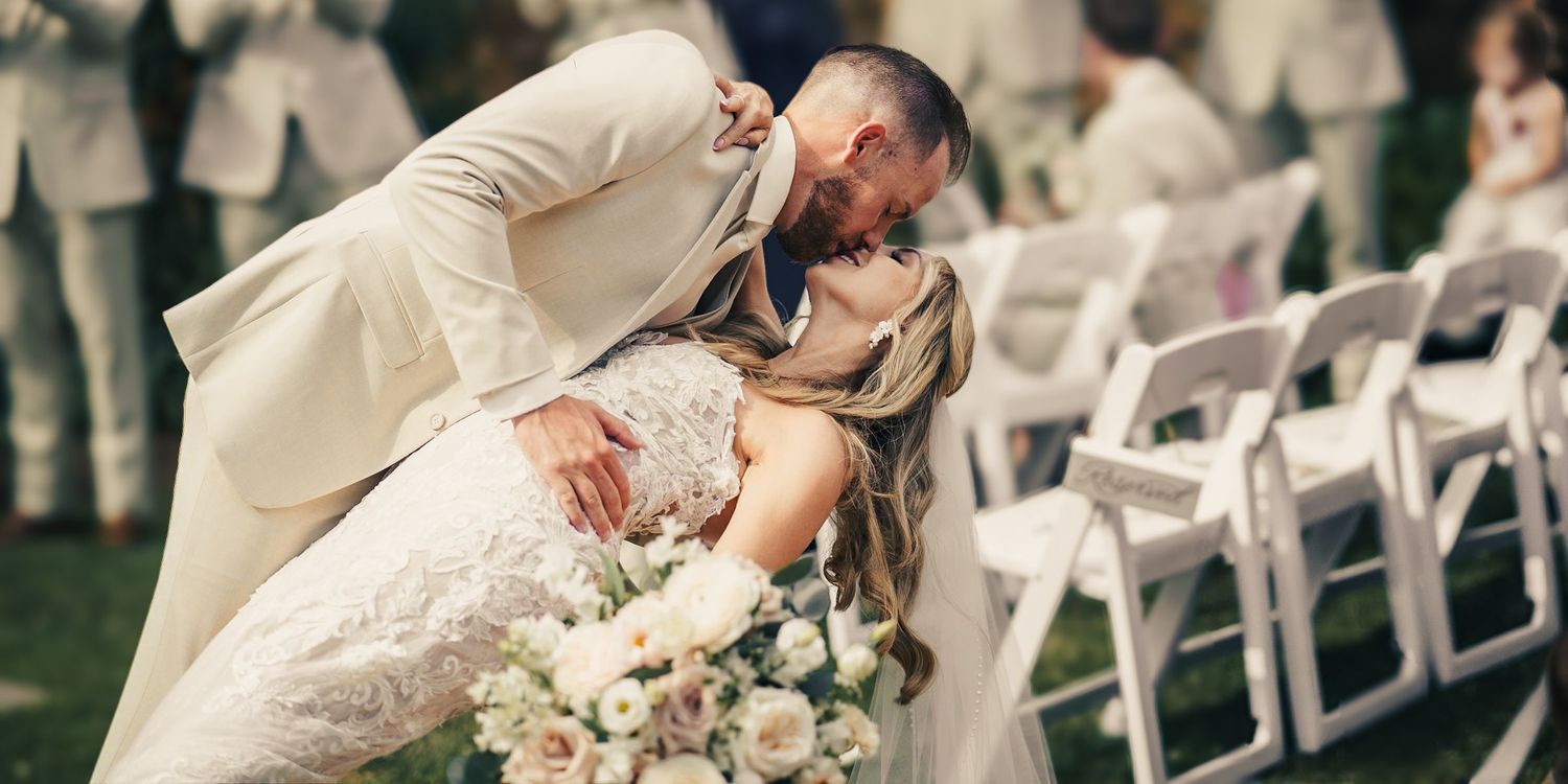 Groom in cream suit dips bride in lace wedding dress for a romantic kiss outdoors at elegant garden ceremony.