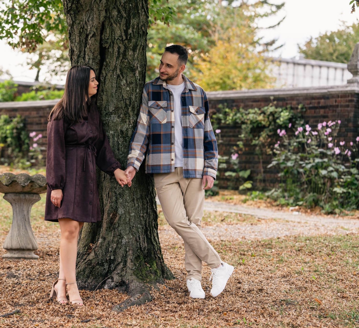 A couple stands together beneath a large tree in a park during autumn with fallen leaves scattered on the ground.