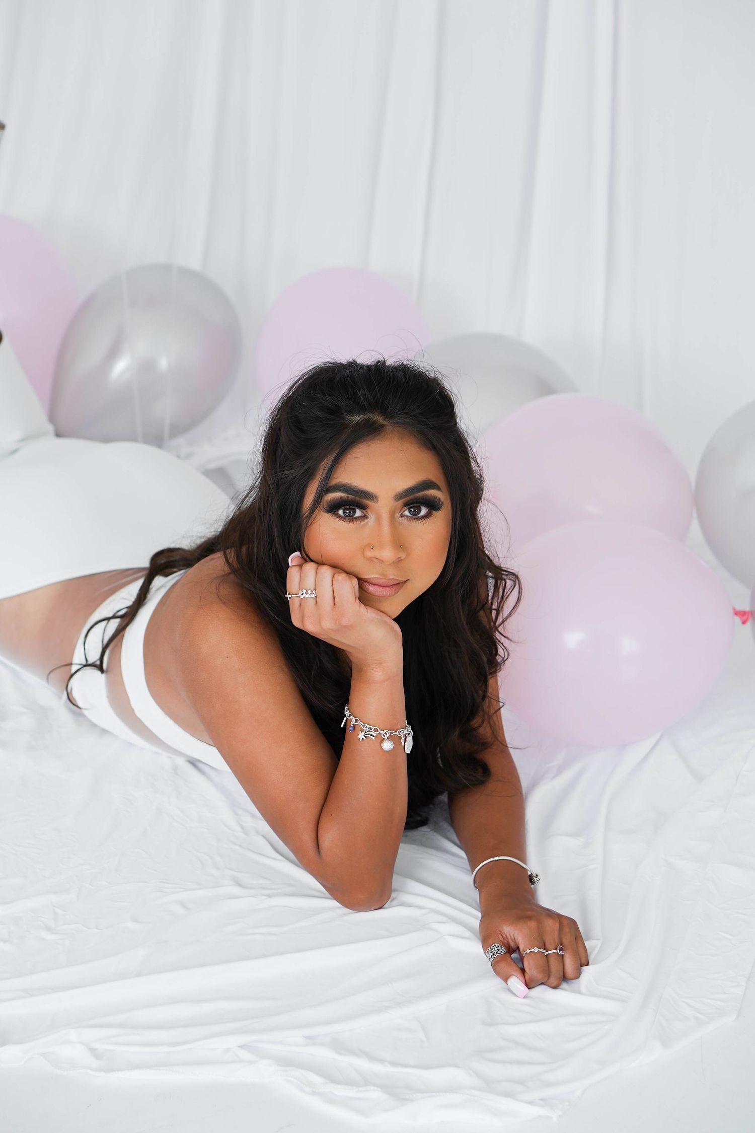 Empowering confident woman in white dress, laying in a white minimalist studio with pink and gray balloons.