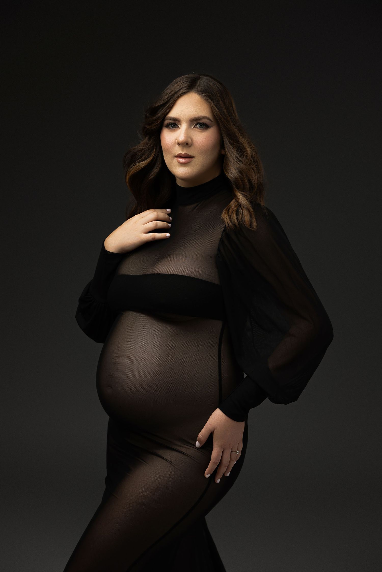 Fashion model poses in sheer black long sleeve bodysuit against dark background in dramatic studio lighting.
