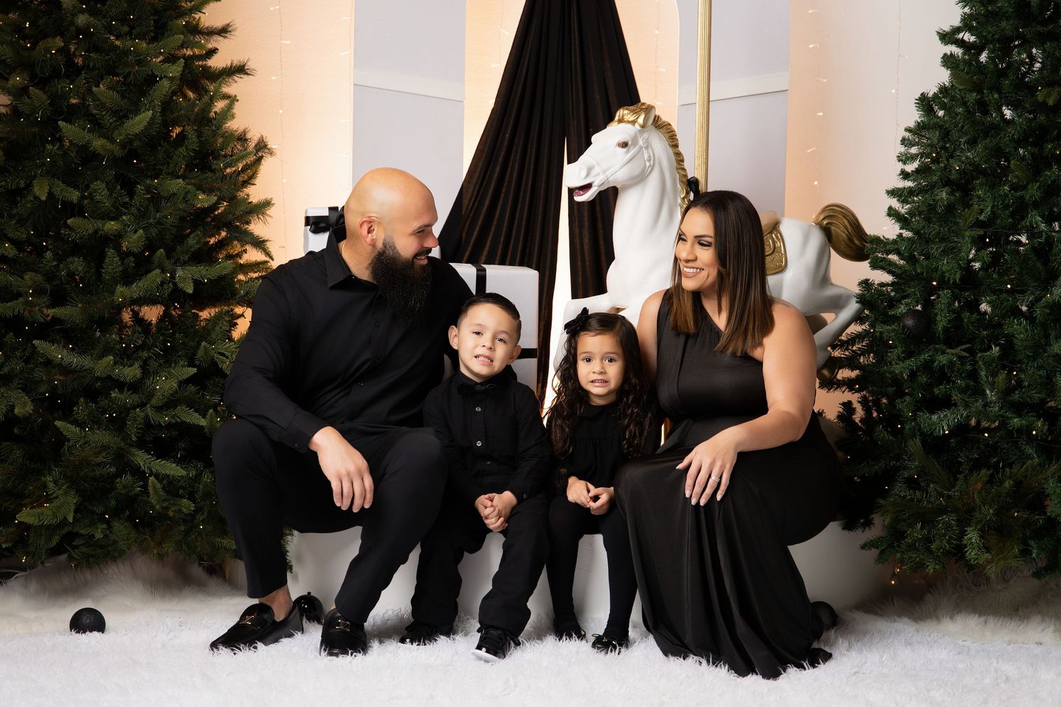 A family in matching black outfits poses on a snowy holiday set with Christmas trees and a white carousel horse behind them.