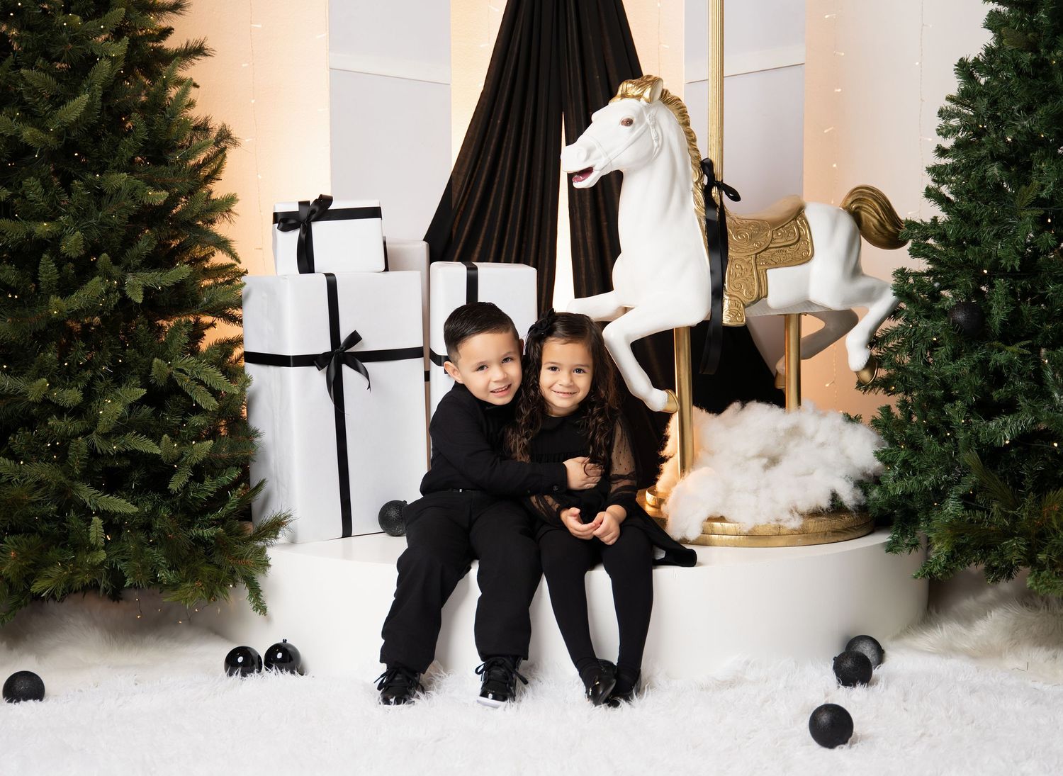 Two children in black outfits sit together on a white platform beside a carousel horse, gift boxes and Christmas trees in a snowy holiday sc