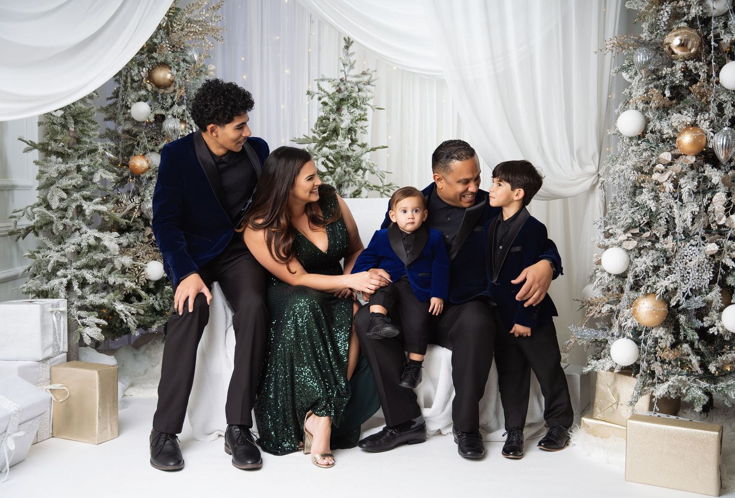 Family of five poses joyfully among decorated Christmas trees and gifts in elegant holiday attire.