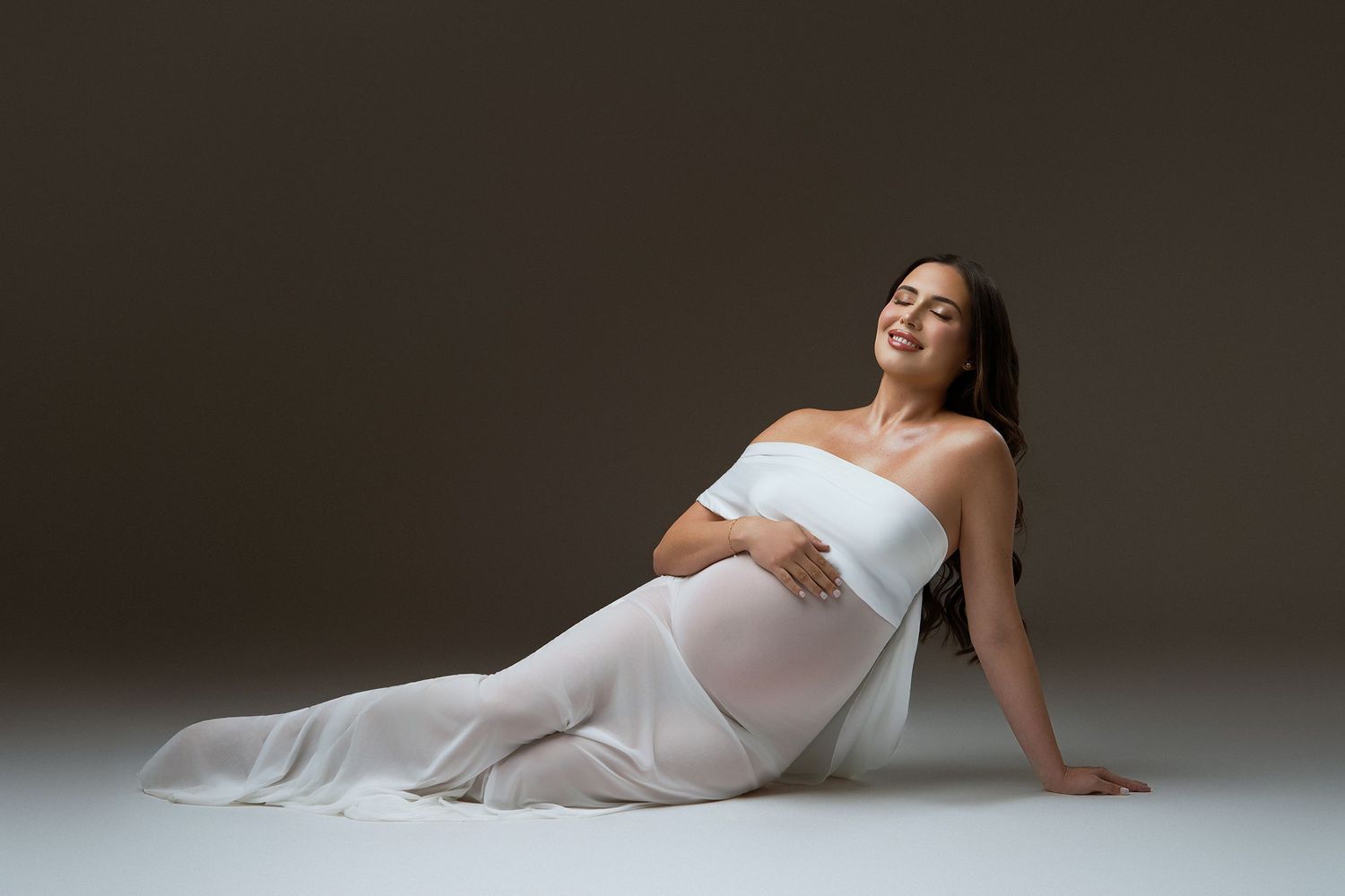 Pregnant woman in white strapless gown sitting gracefully on floor, cradling baby bump, smiling softly against dark background.
