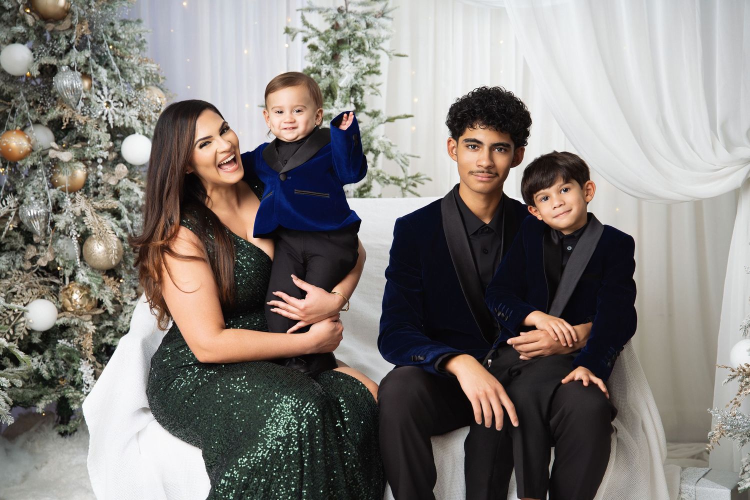 Family of four posing for a festive Christmas photo near a decorated tree in elegant holiday attire.