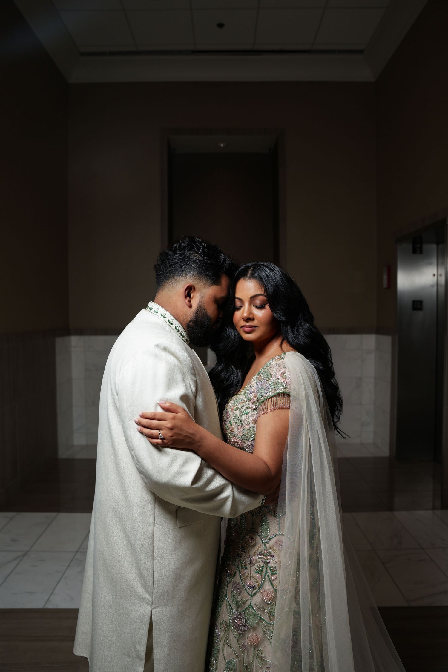 A romantic moment between a couple in traditional Indian wedding attire standing in a dimly lit hallway.