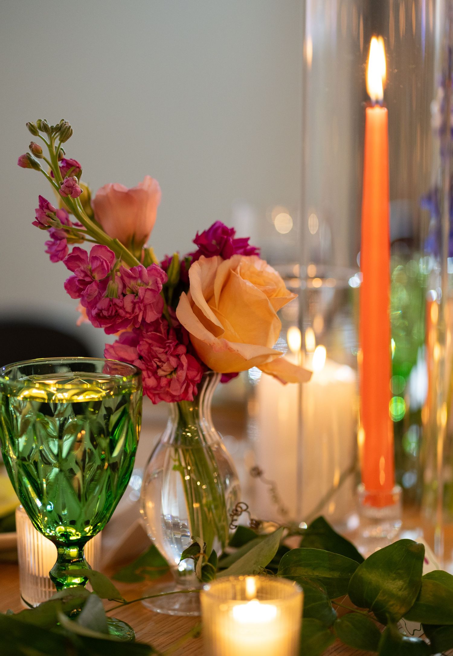 Elegant table setting with lit orange candles, green glass vases, and pink roses alongside purple flowers in soft lighting in Chicago