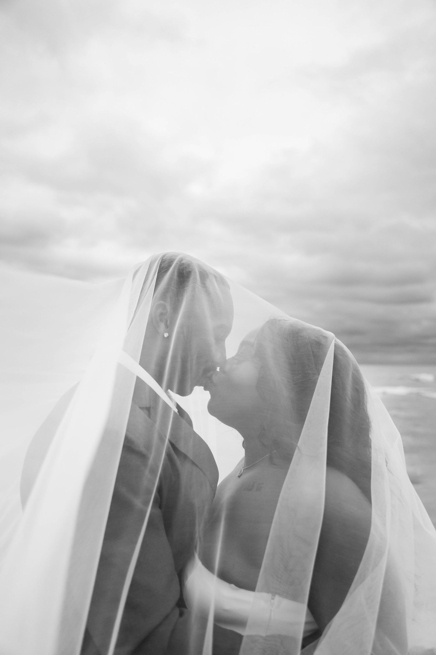 A romantic black and white wedding photo showing a couple sharing a kiss under a flowing sheer veil.