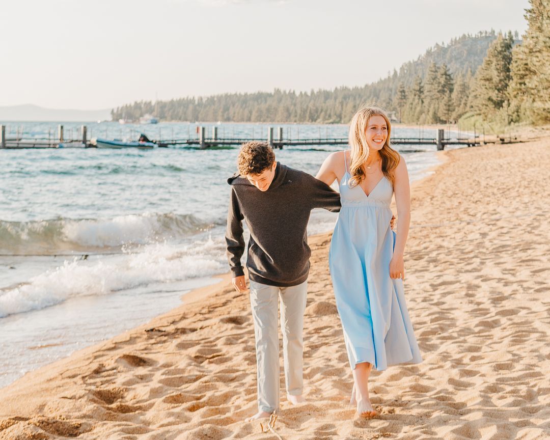 A couple walks hand in hand along a sandy beach with mountains and trees in the background during golden hour.