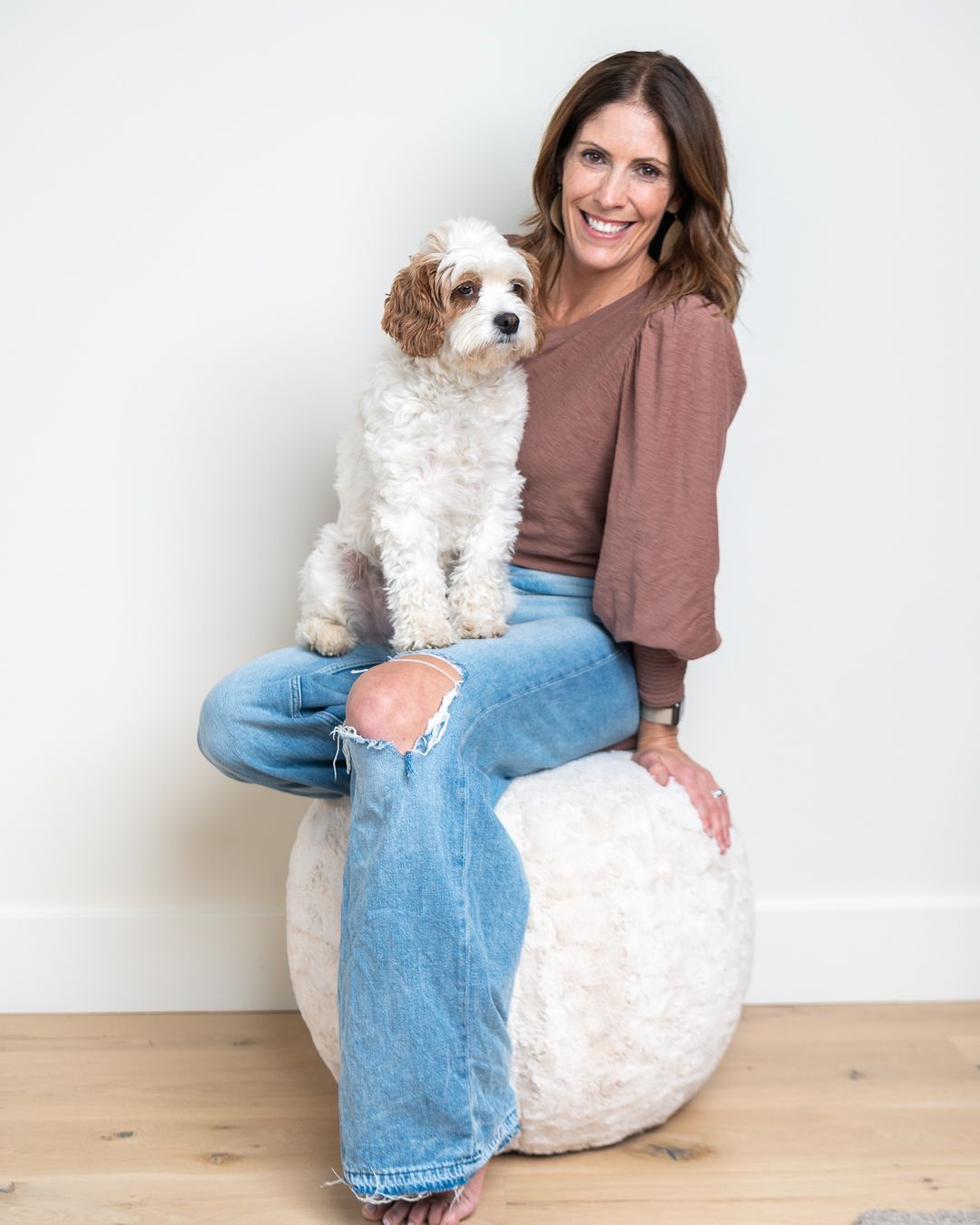 Someone in distressed jeans and a brown top sits on a white ottoman with their small fluffy white dog.
