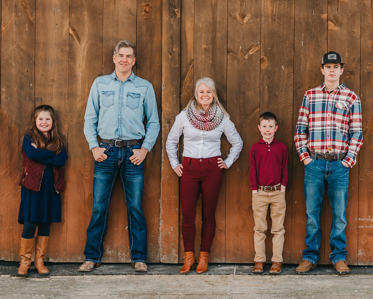 Family of five wearing coordinated fall outfits standing against rustic wooden barn wall.