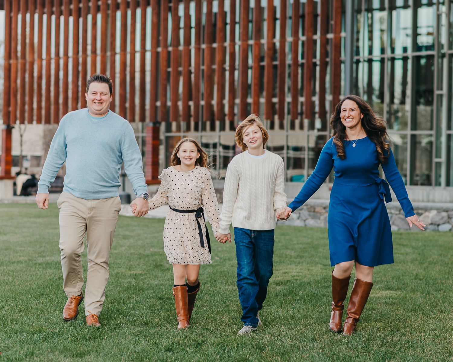 Family of four walking together and holding hands on green grass in front of a modern building exterior.