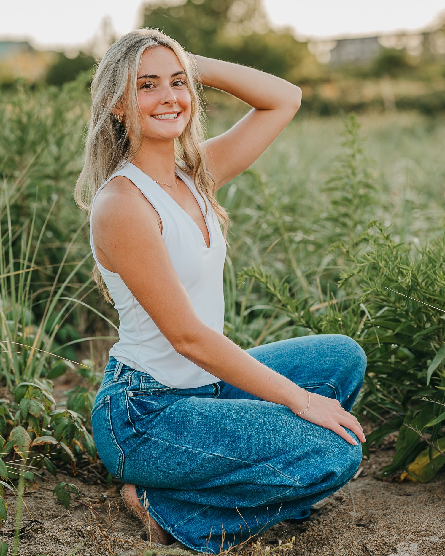 Person in white tank top and blue jeans poses outdoors in a garden setting during golden hour.