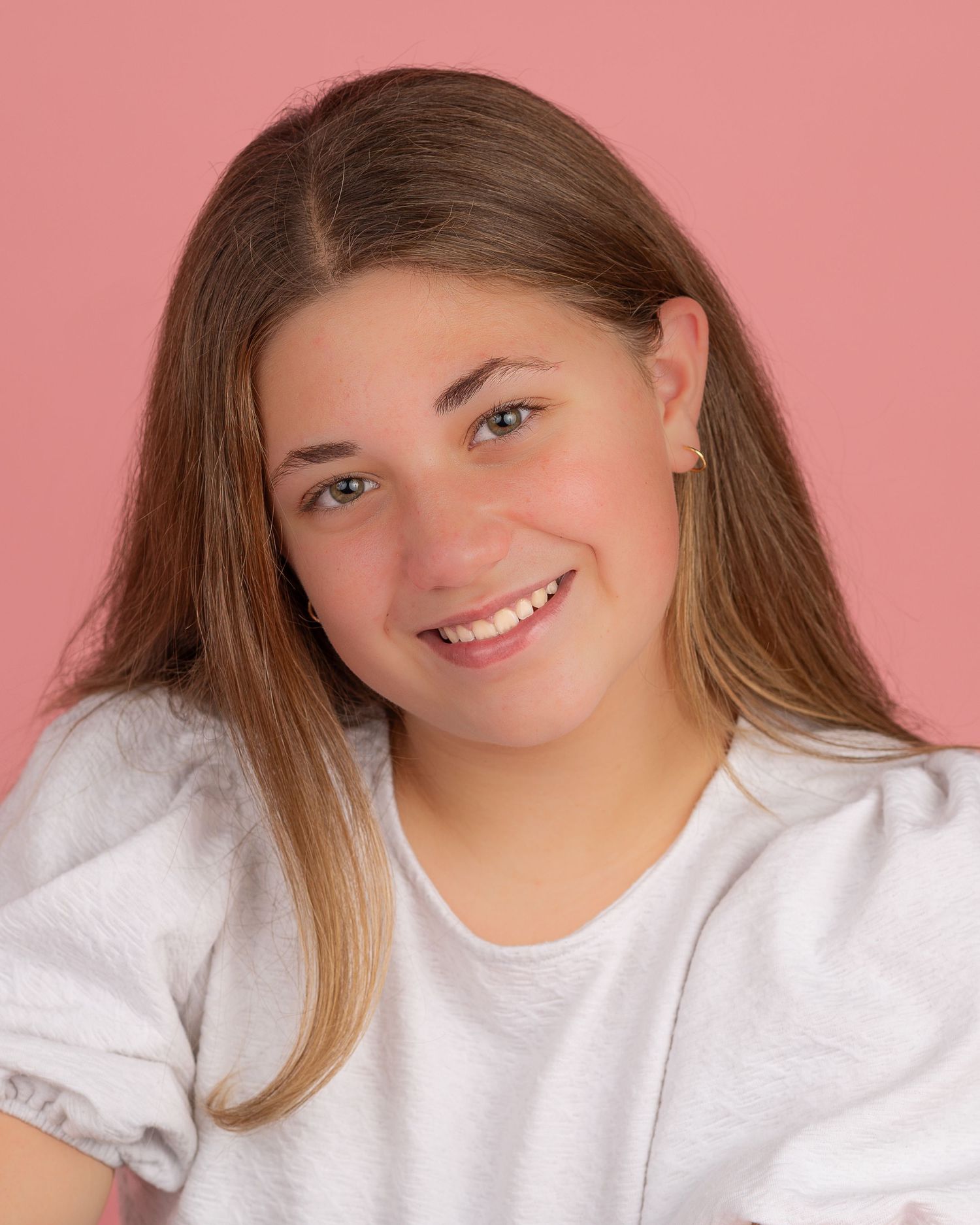 Portrait photo against pink backdrop showing a smiling person in a white top with long brown hair.