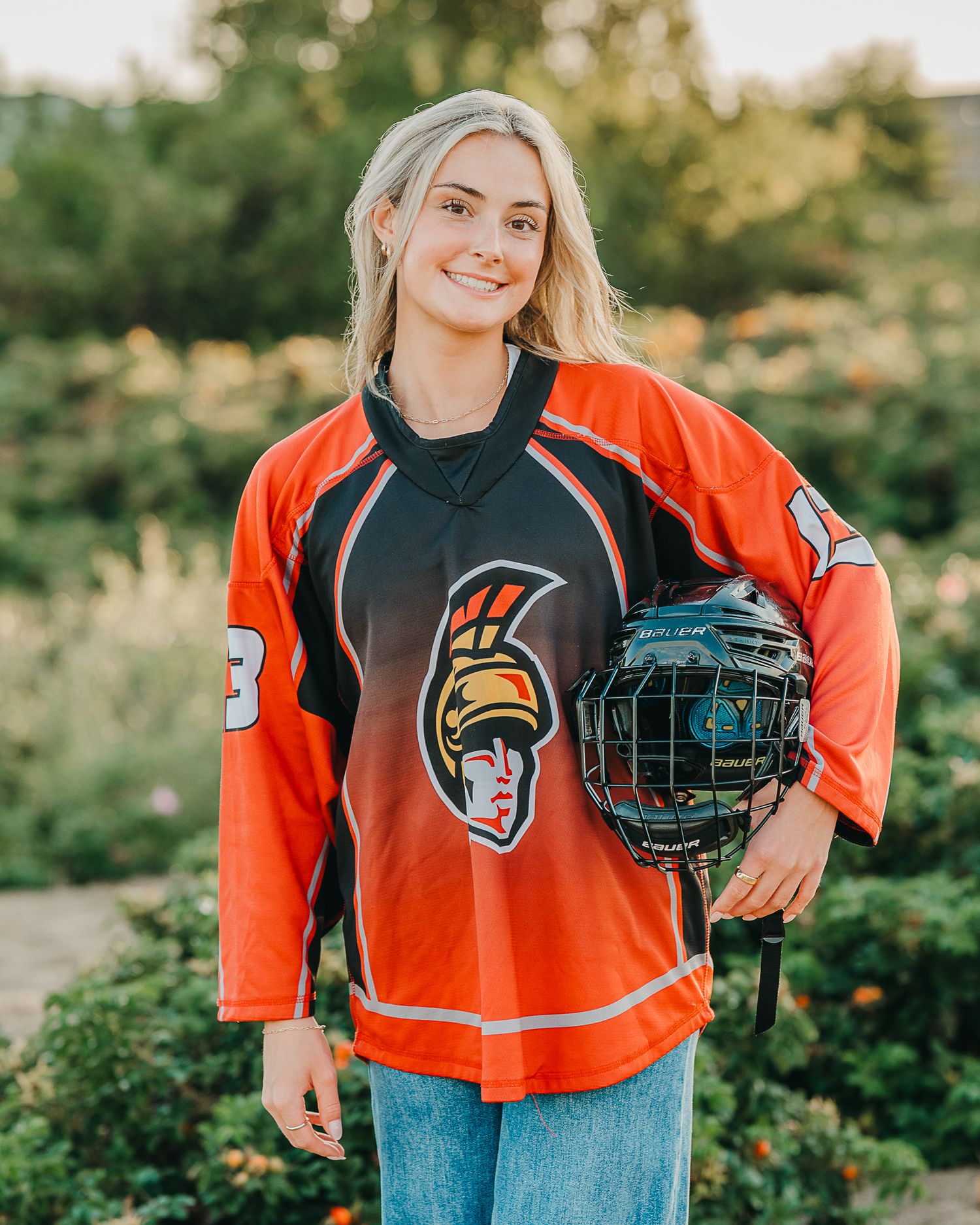 Person wearing an orange and black sports jersey poses with hockey equipment in a sunny outdoor setting.