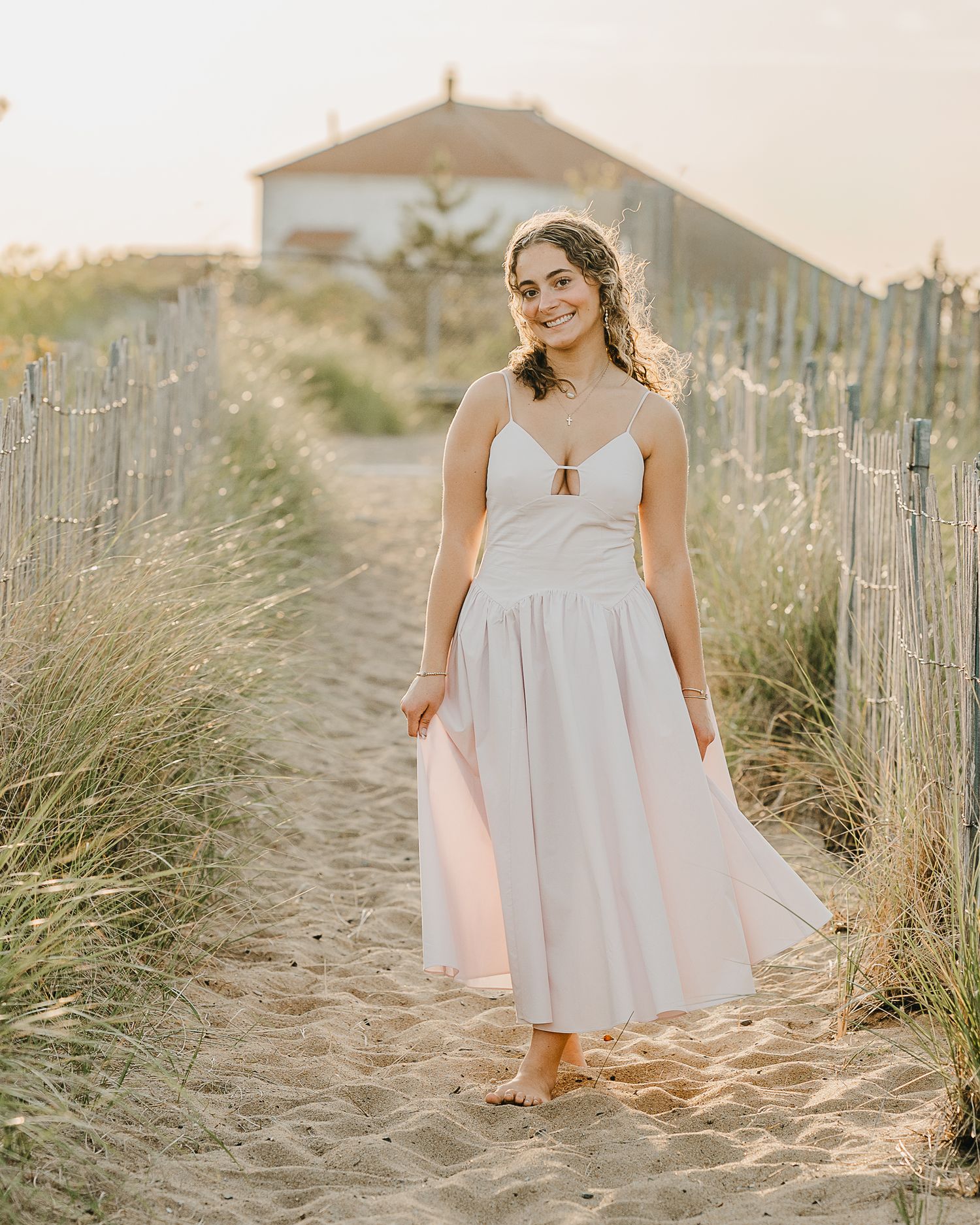 A person in a flowing white dress walks along a sandy beach path lined with wooden fencing at sunset.