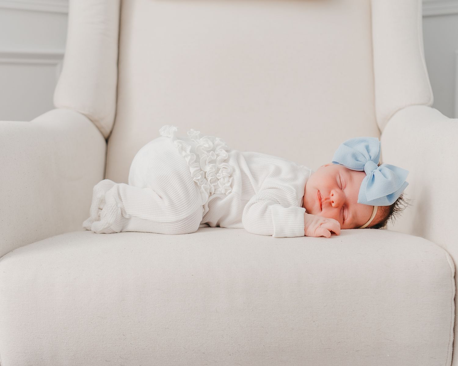 A sleeping newborn in white clothing and blue bow rests peacefully on a cream-colored armchair in Londonderry nh