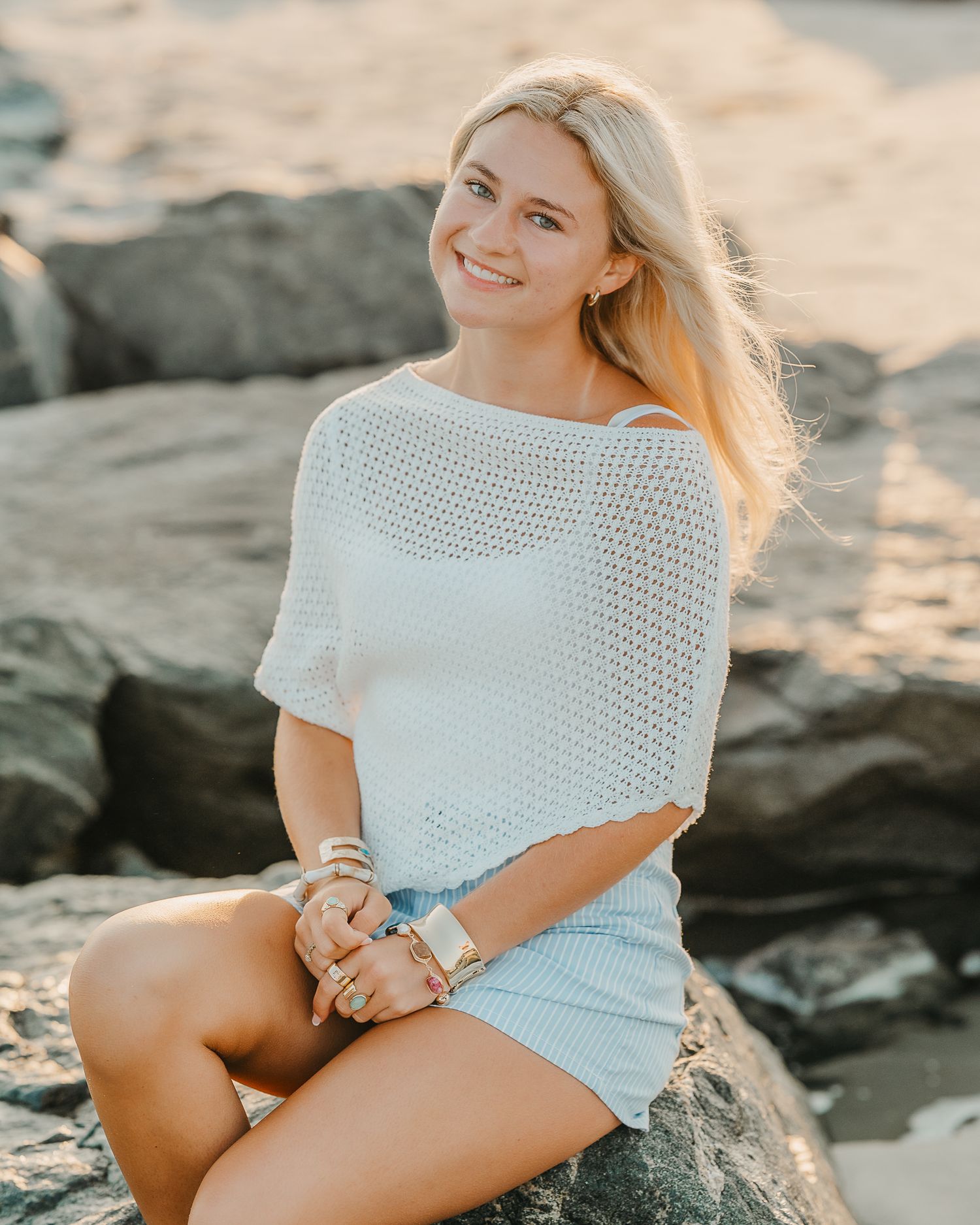 Smiling blonde woman in white knit top and denim shorts sitting on rocks at a sunny beach.