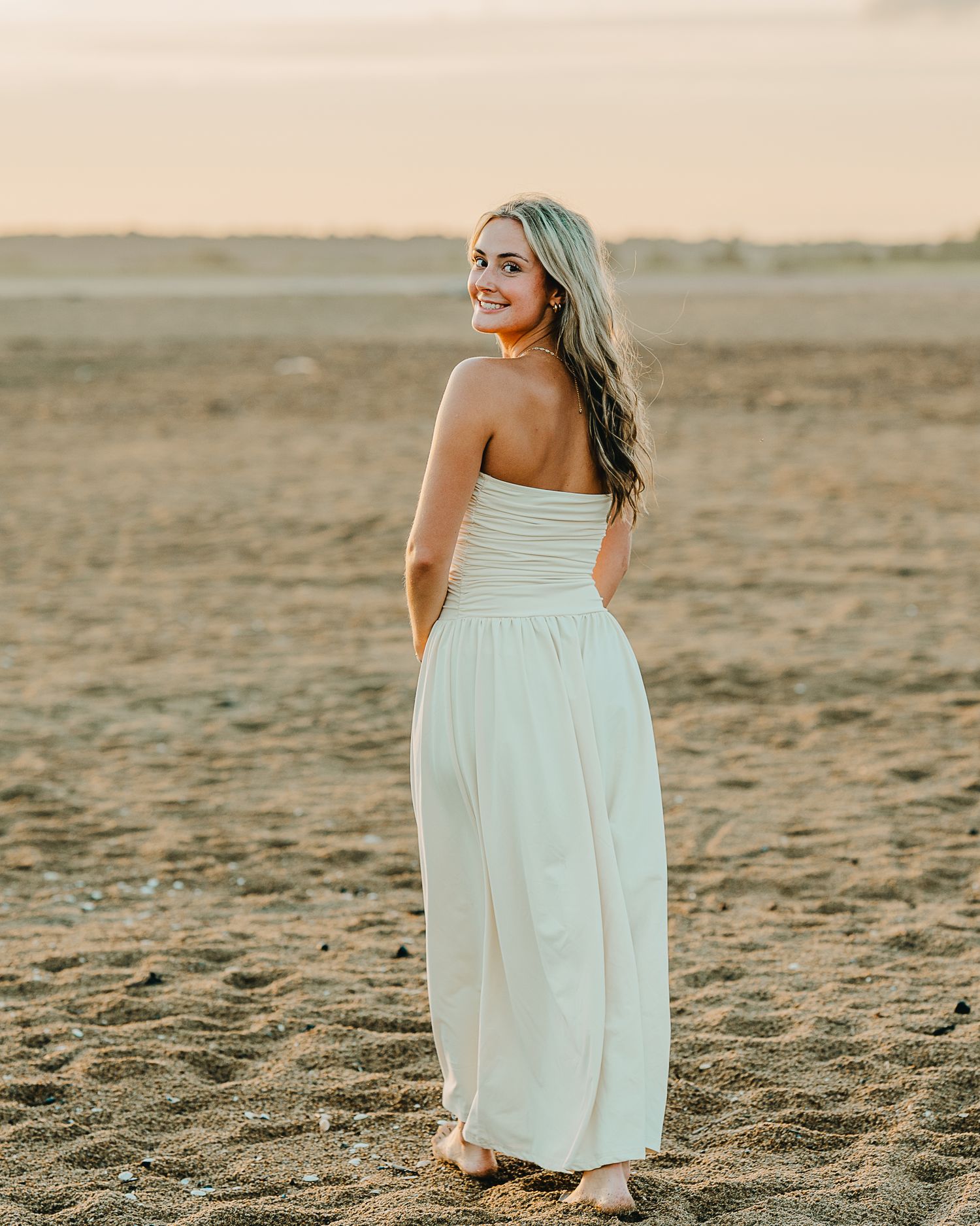 Woman in a white sundress walking barefoot on a sandy beach at sunset.