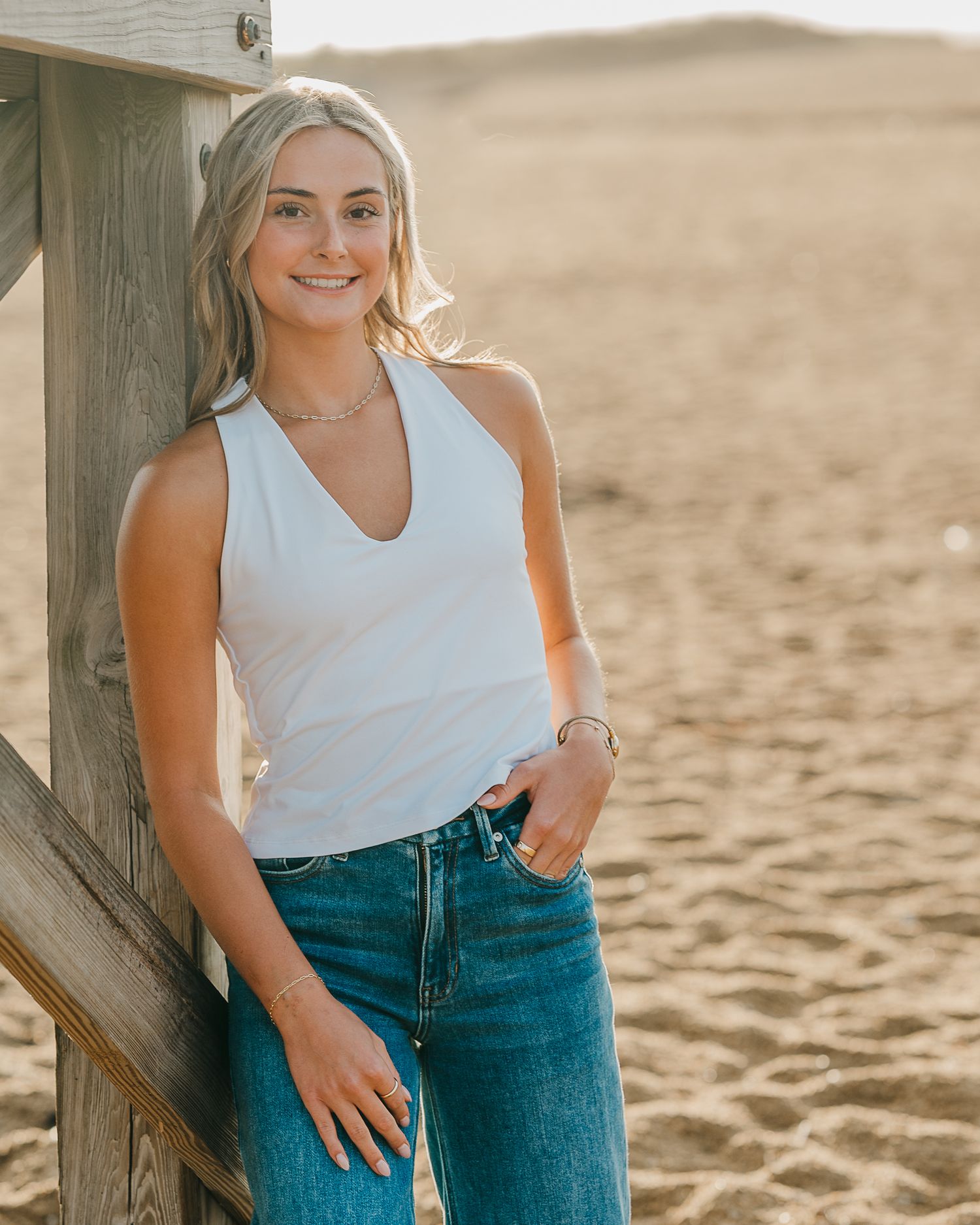 Person in a white tank top and blue jeans poses casually against a wooden railing at the beach during sunset.
