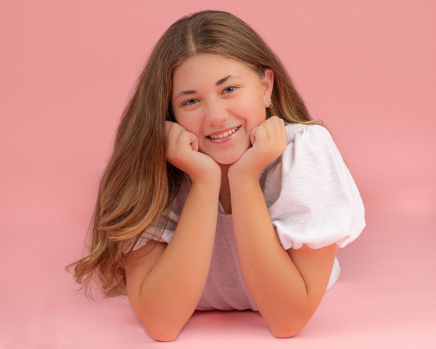 A teen with long brown hair wears a white puff-sleeve top while posing for a headshot casually against a pink background.