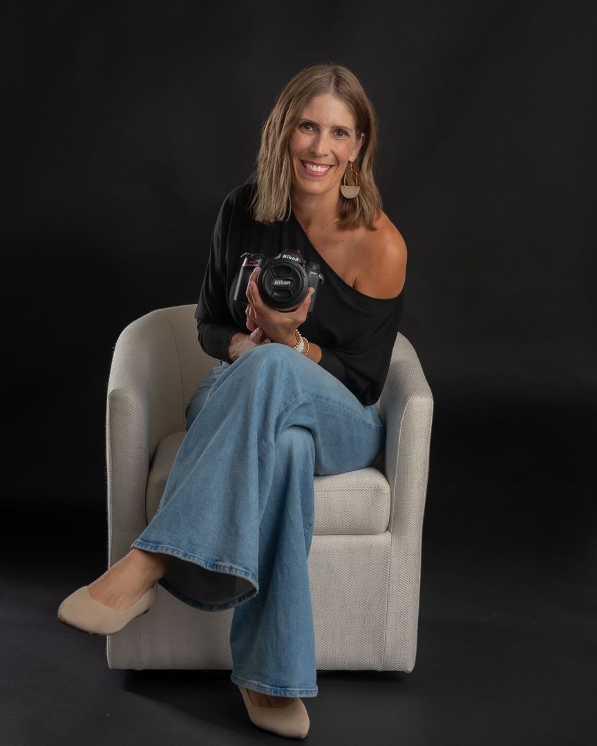 Smiling woman sitting in white chair holding DSLR camera, wearing black top and jeans, professional photographer portrait.