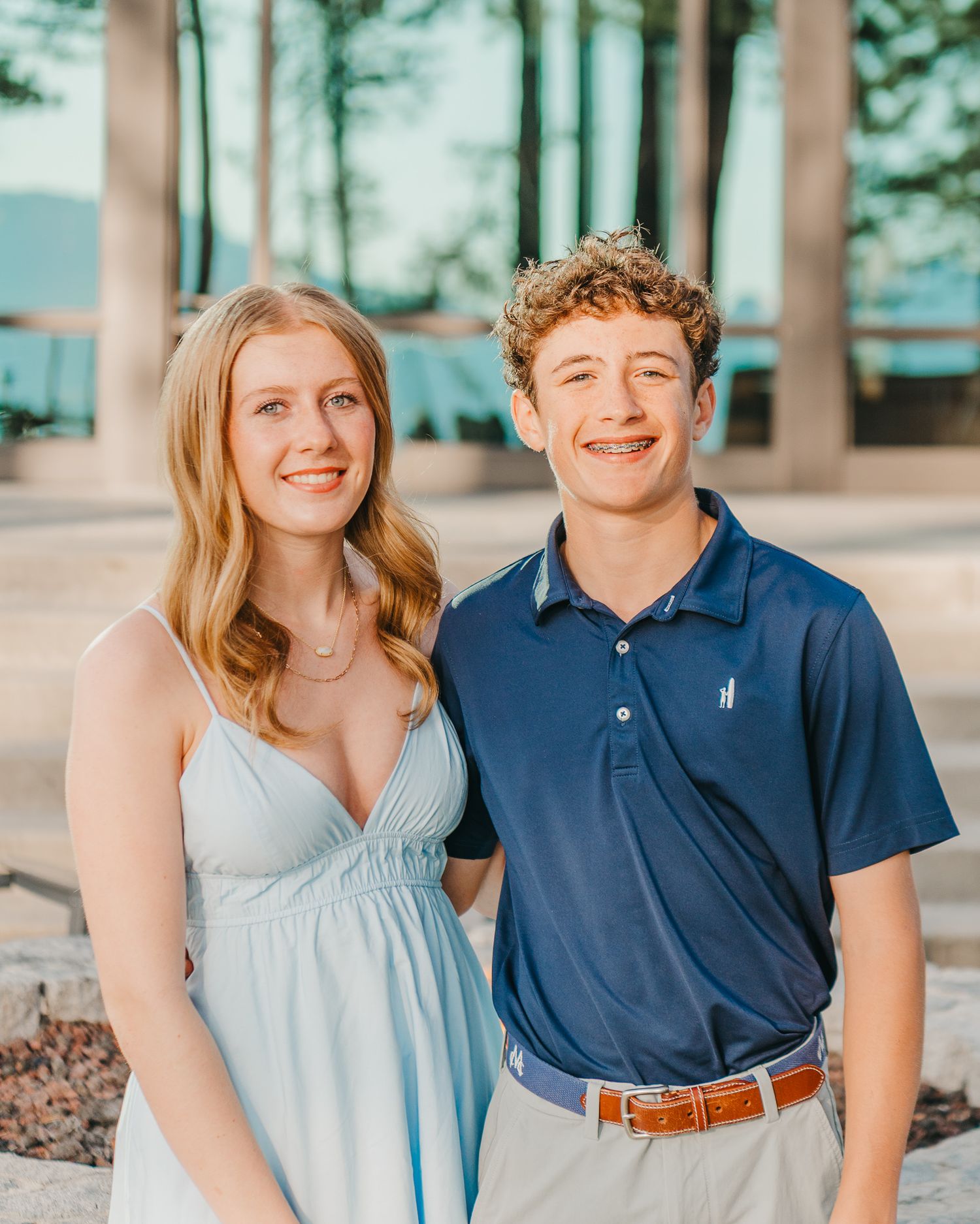 A smiling young couple poses outdoors, woman in light blue dress and man in navy polo shirt with braces.