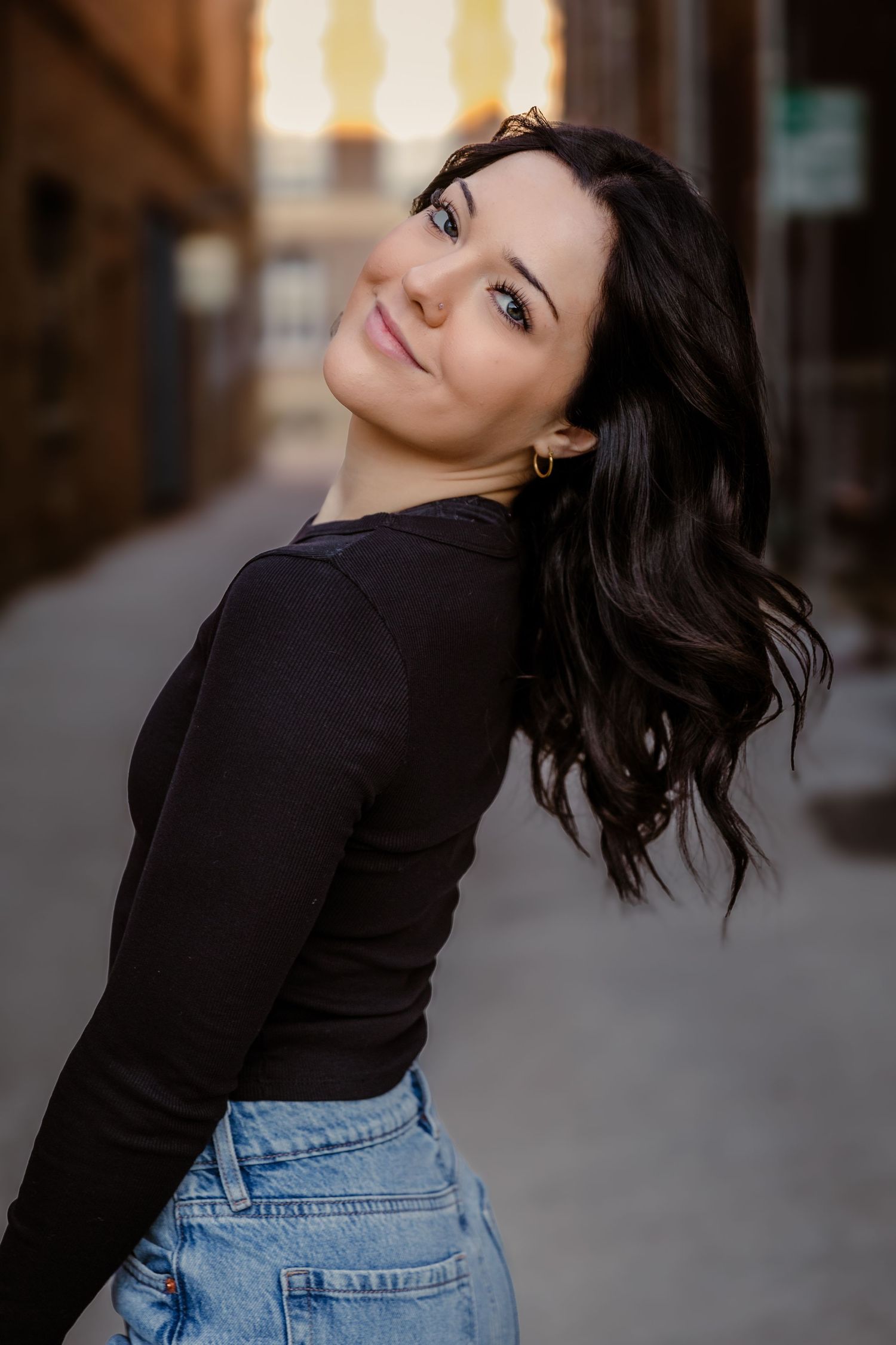 Woman in black sweater and jeans poses casually against urban backdrop during sunset photoshoot.