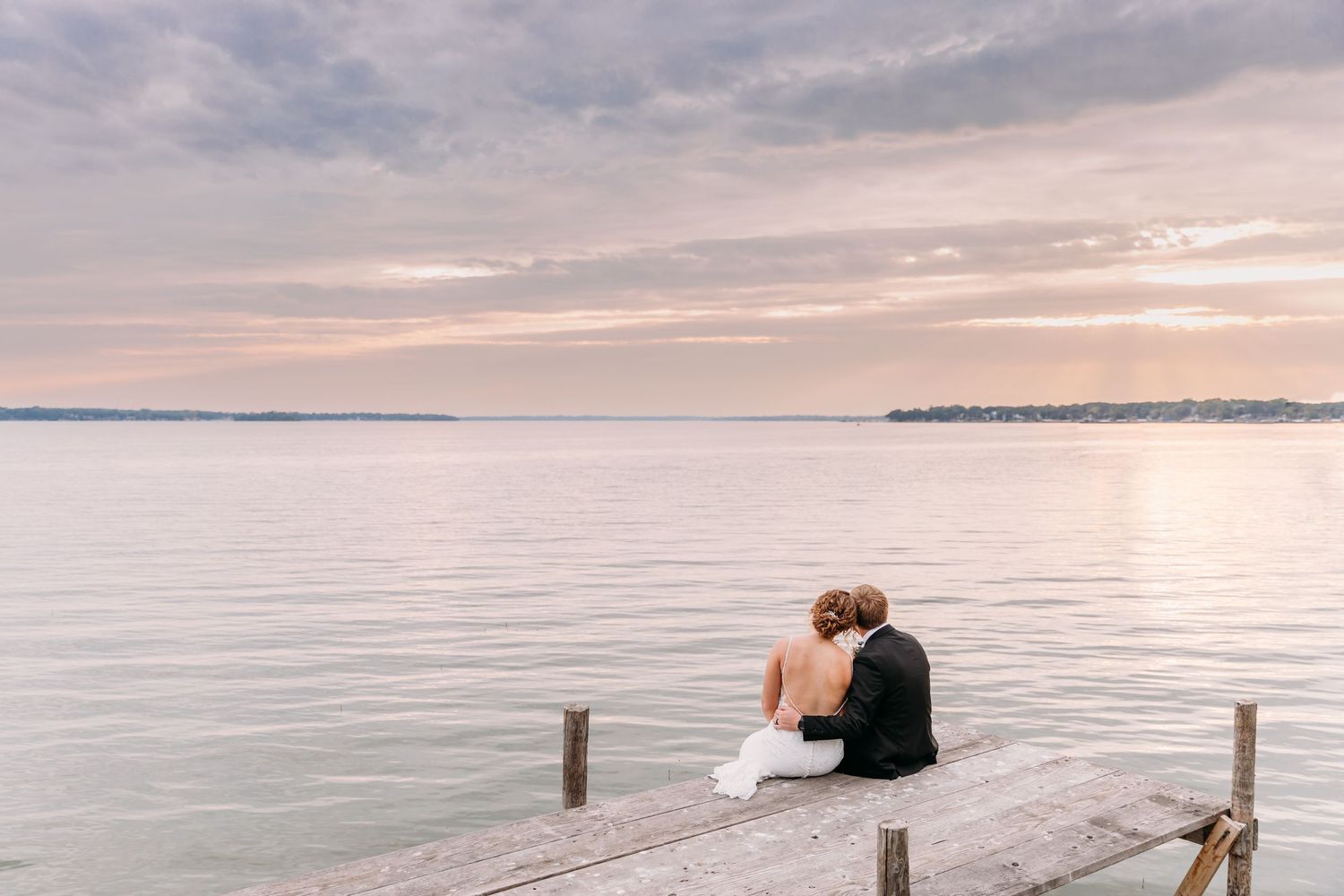 Couple sits together on wooden dock overlooking calm lake during pastel sunset.