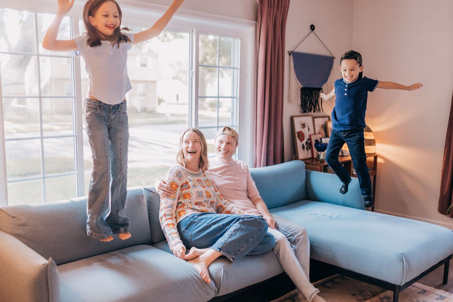 Playful family moment as children jump on blue couch while parent watches from below.