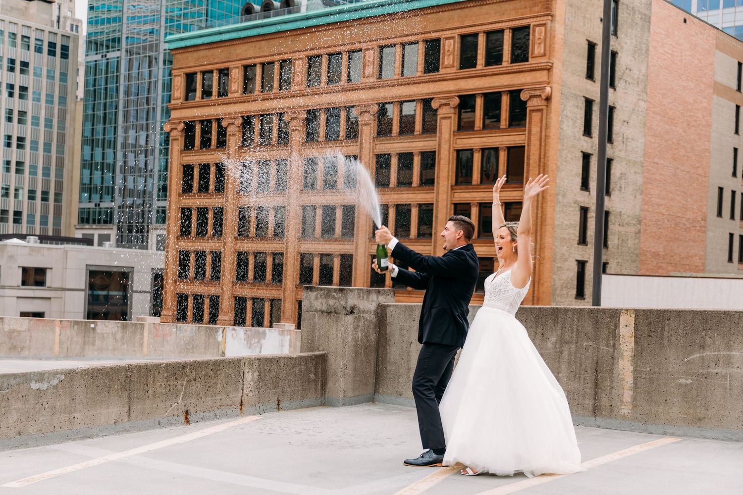 A couple in wedding attire celebrates together on an urban rooftop with brick buildings in the background with a champagne spray.