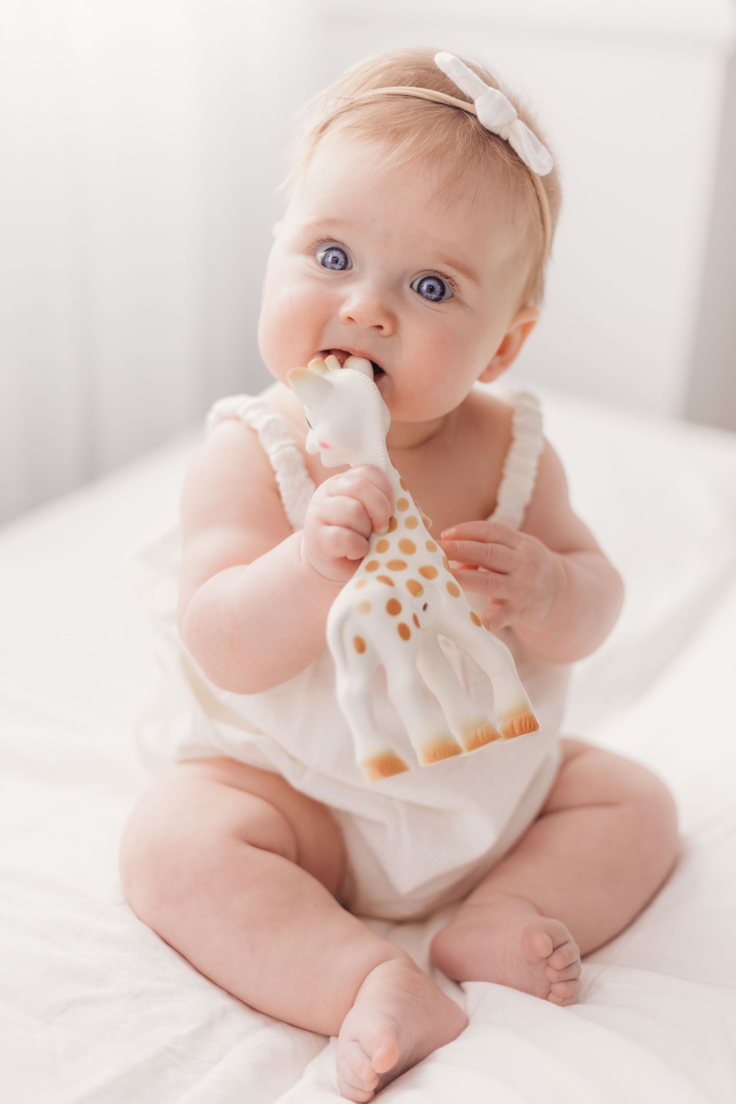 Baby in white outfit chews on Sophie the Giraffe teether toy while wearing bow headband.