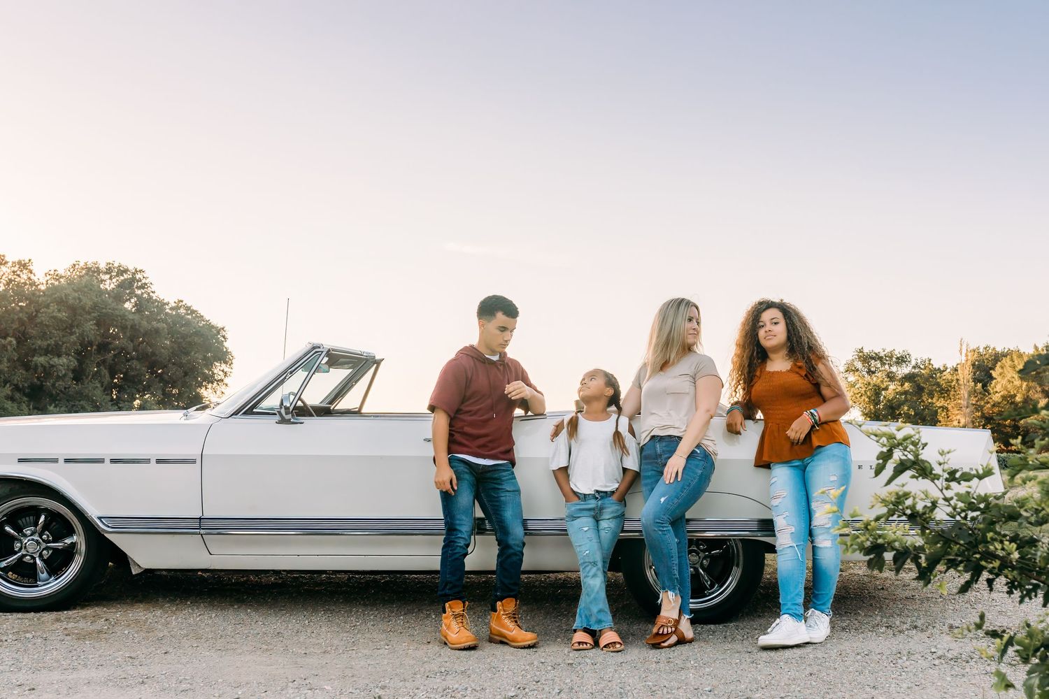 Family of four poses together next to classic white car during sunset photoshoot.
