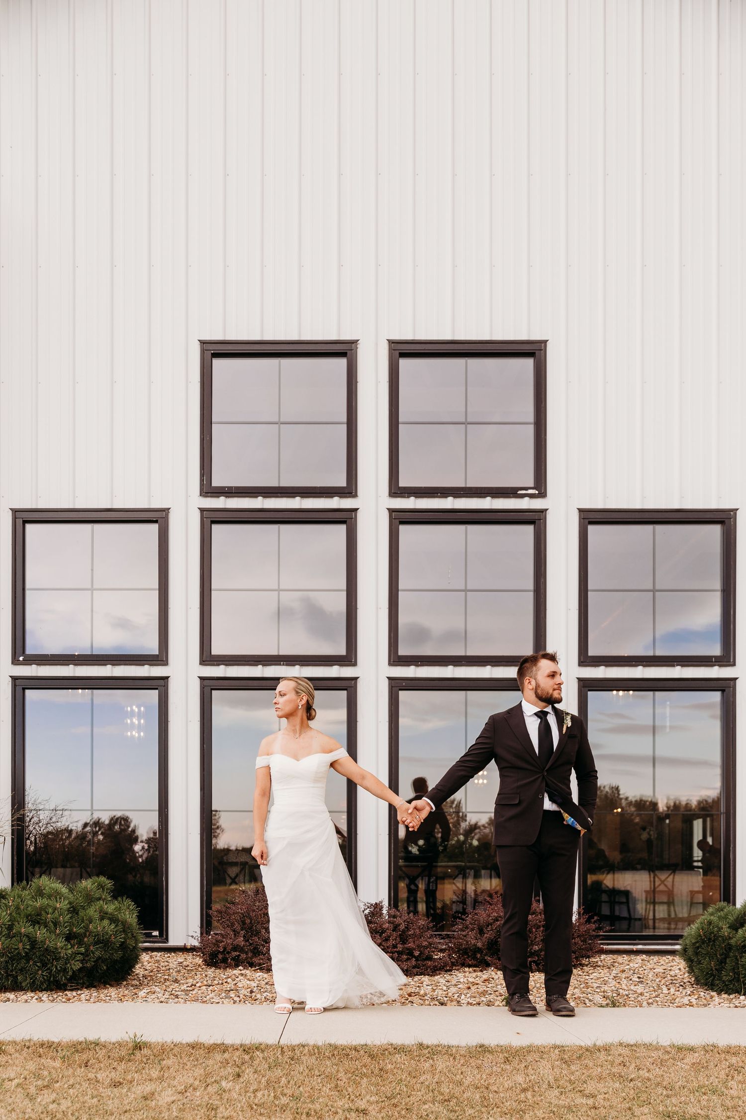 A couple in wedding attire playfully runs past a modern building with large windows and grassy landscaping.
