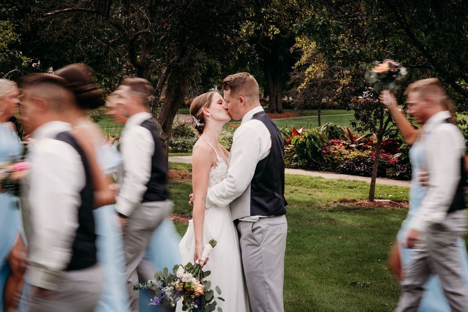 Bride and groom share a kiss while wedding party walks past in a lush garden setting.
