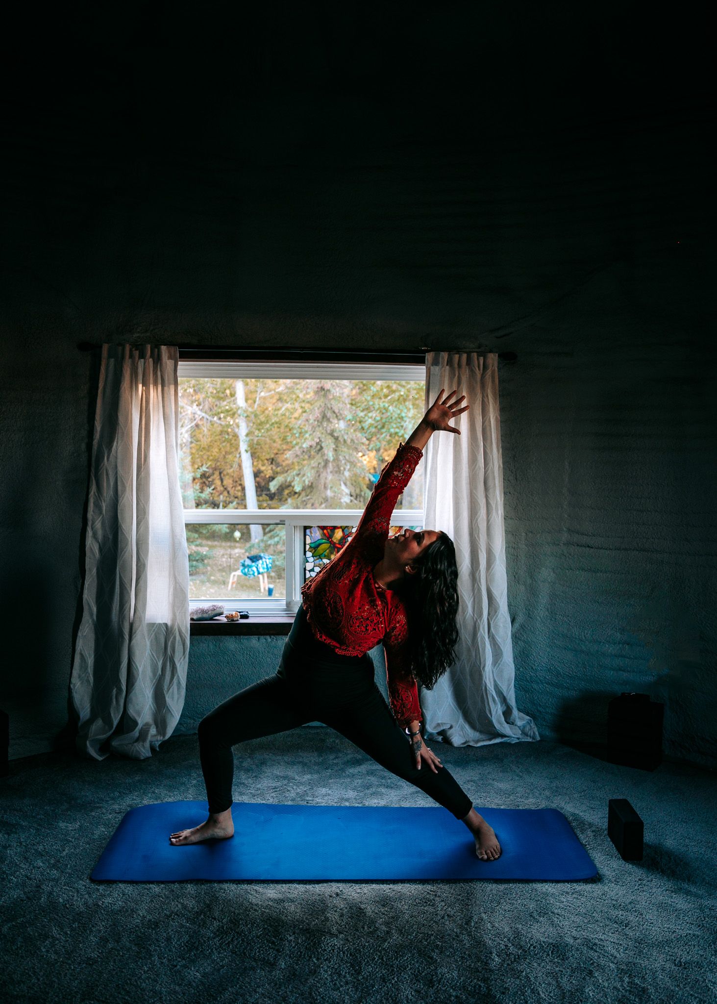A woman practices a warrior yoga pose on a blue mat in a dimly lit room.
