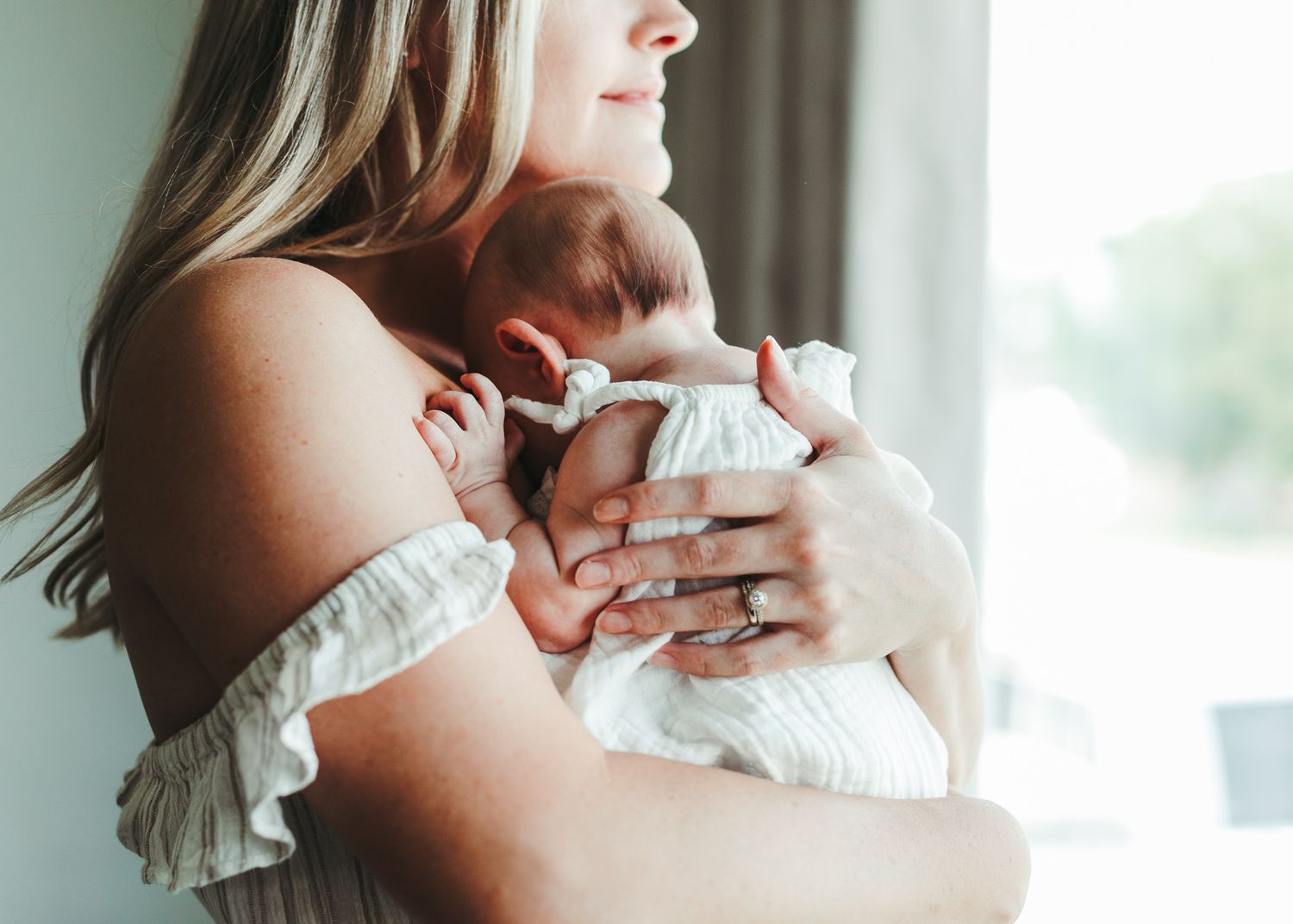 A mother tenderly holds her newborn baby close to her chest near a bright window.