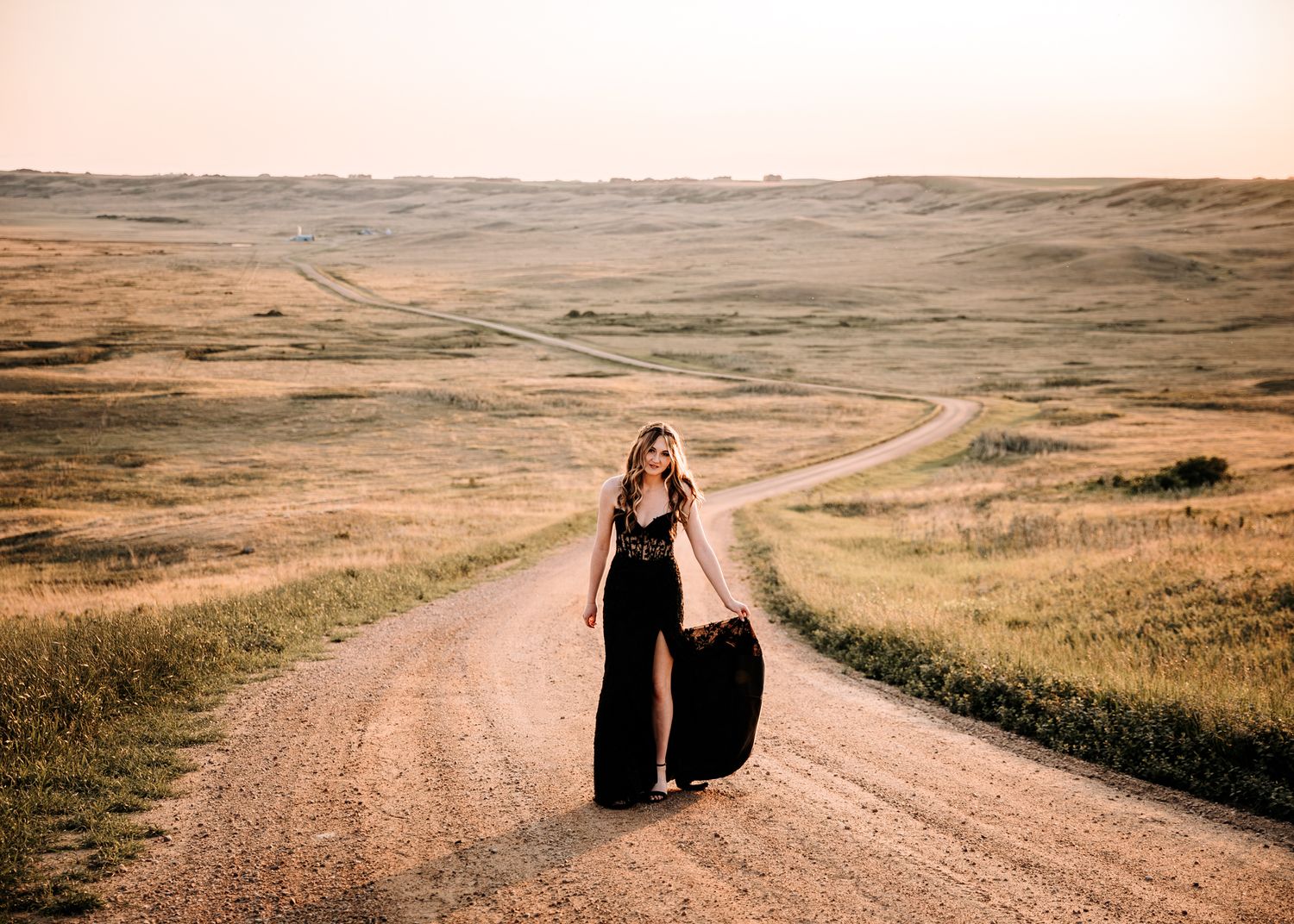 Young woman in elegant black dress walking along a winding dirt road through vast open prairie.
