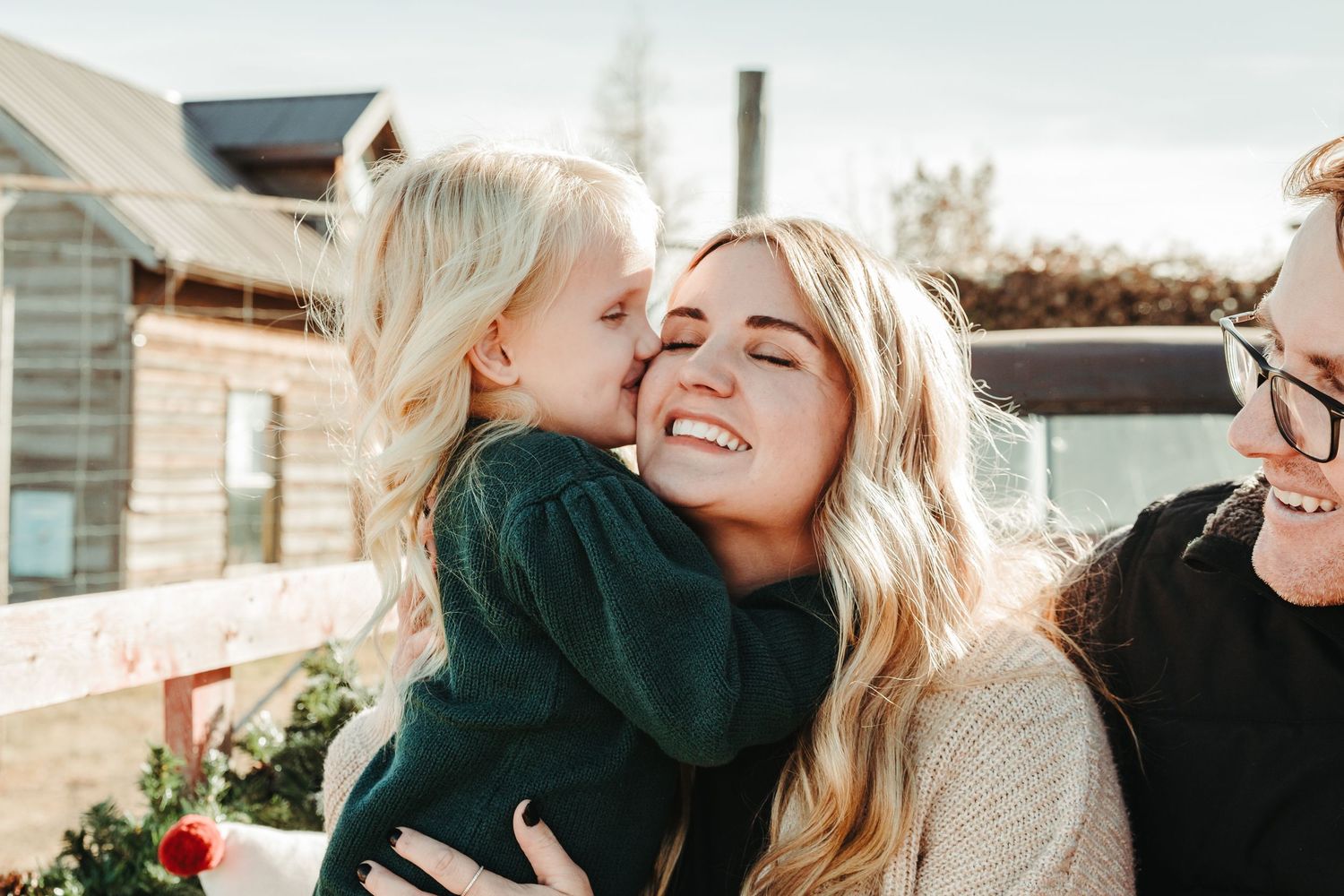 A smiling woman with long blonde hair receives a sweet kiss on the cheek from a young blonde child outdoors.