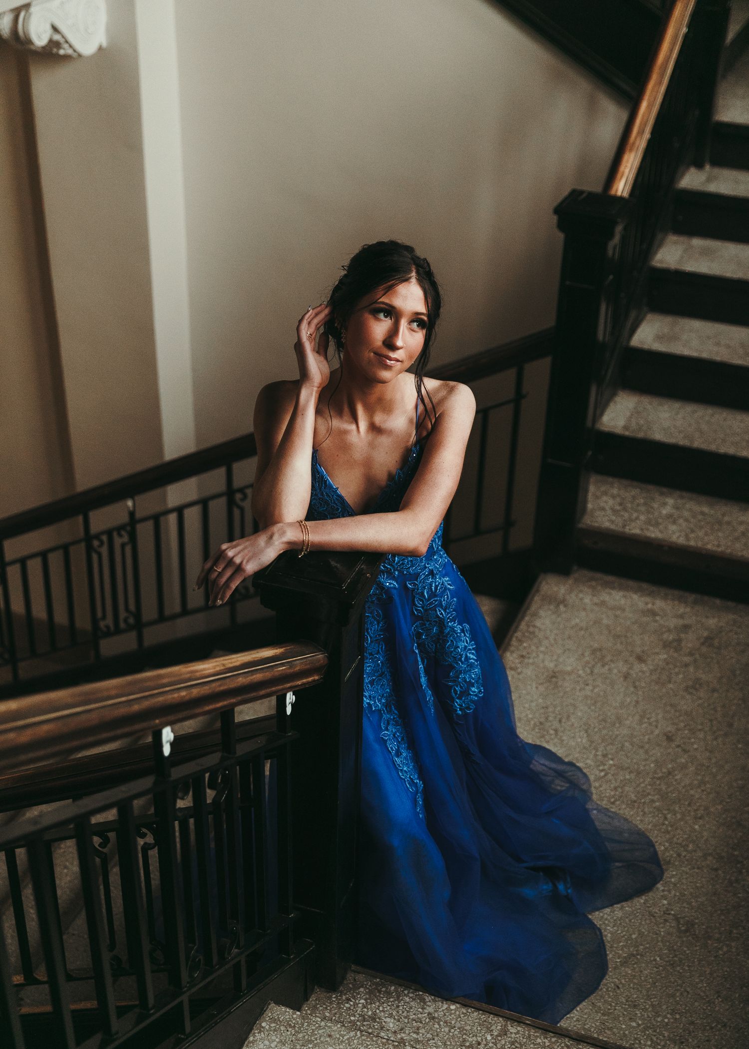 Elegant young woman in a blue lace gown posing gracefully on a staircase in a classic interior. Senior grad photos. Graduation photos.