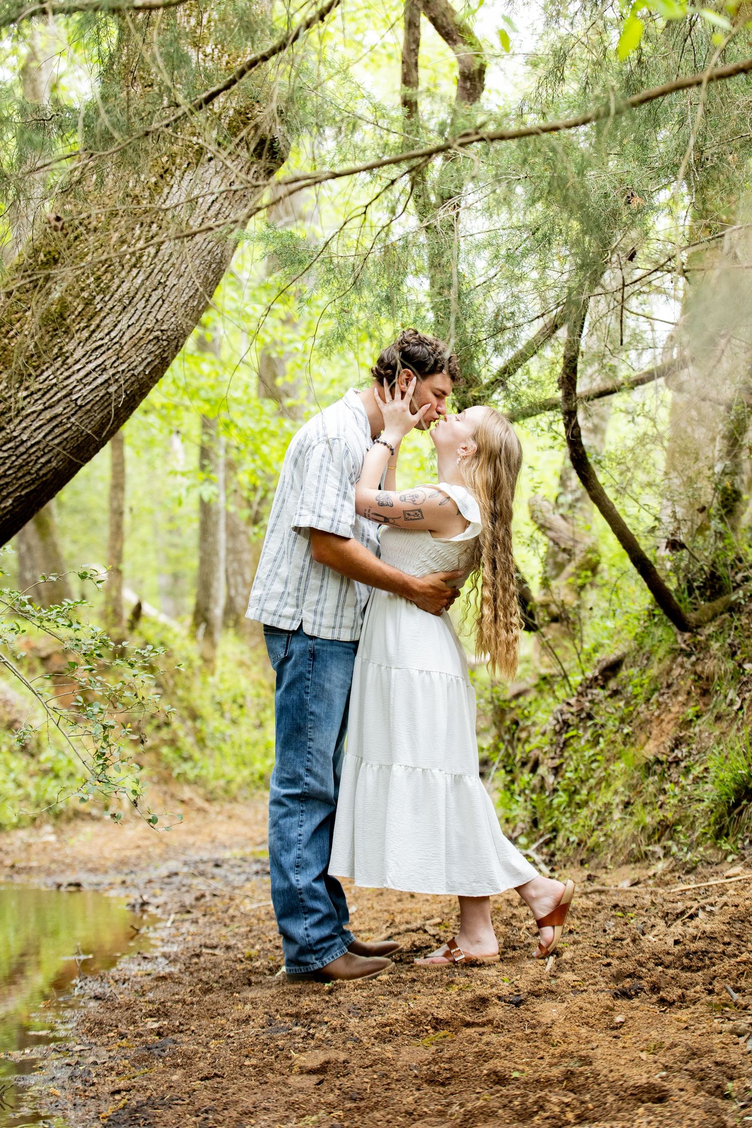 Couple sharing a romantic kiss on a forest path surrounded by lush green trees and a small stream.