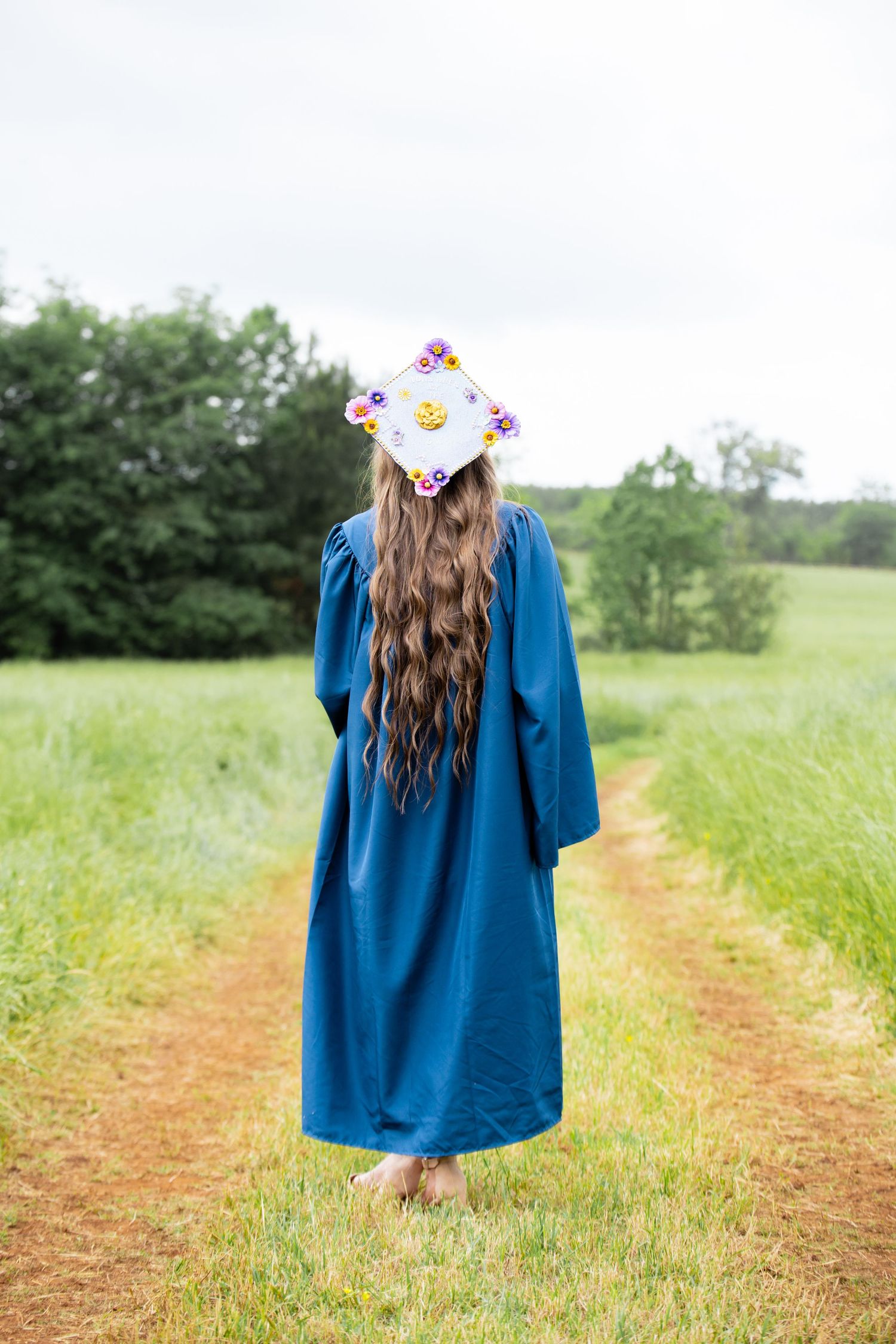 Graduate in blue cap and gown with floral mortarboard stands on dirt path surrounded by green fields.