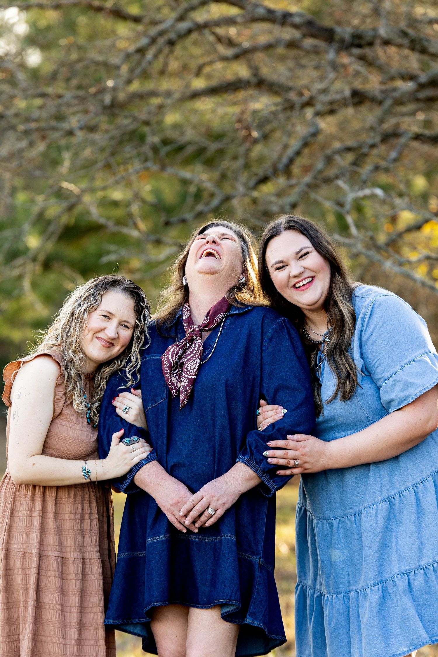 Three women laughing and embracing outdoors in colorful dresses among autumn trees.