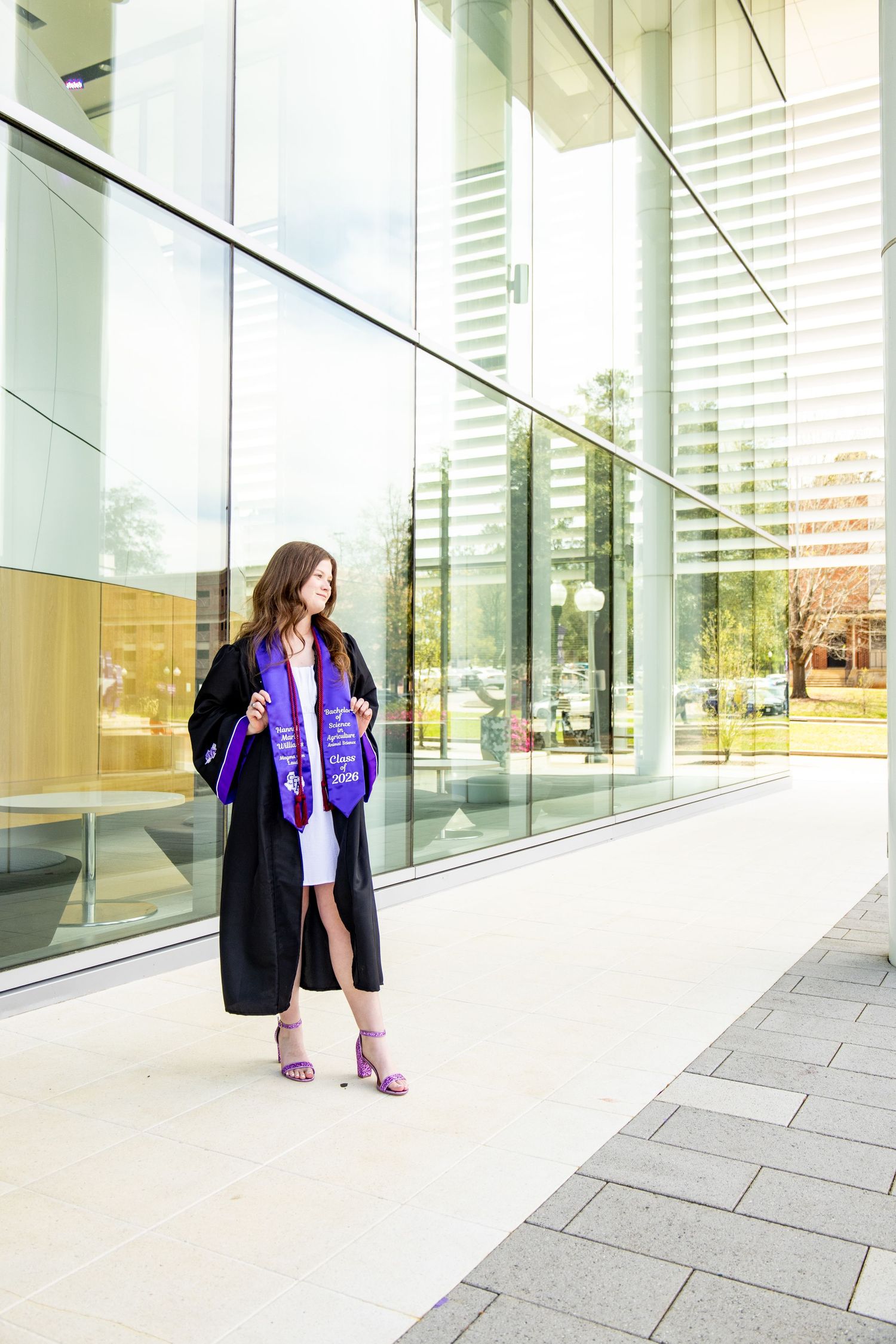 Graduate in black gown and purple stole poses confidently outside a modern glass building on a sunny day.