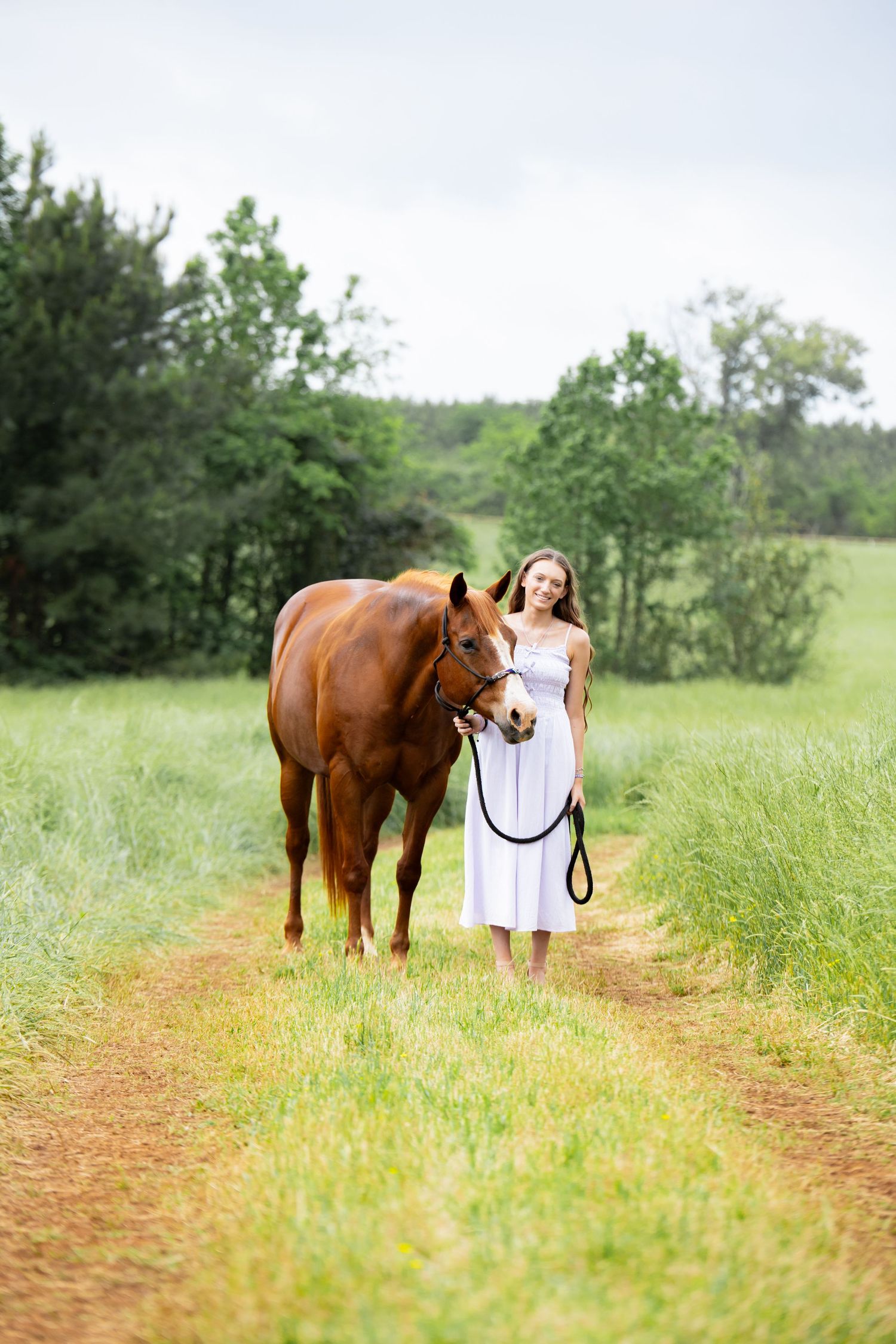 Young woman in white dress leading a chestnut horse along a rural dirt path surrounded by lush green fields.