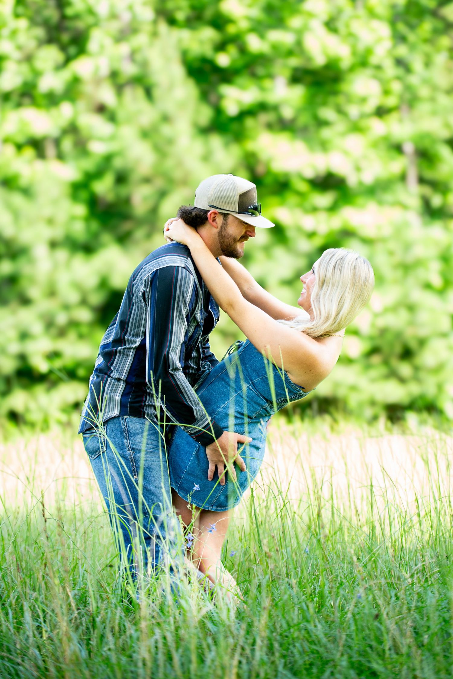 Couple embracing in tall green grass outdoors, man in plaid shirt and cap lifting woman with blonde hair.