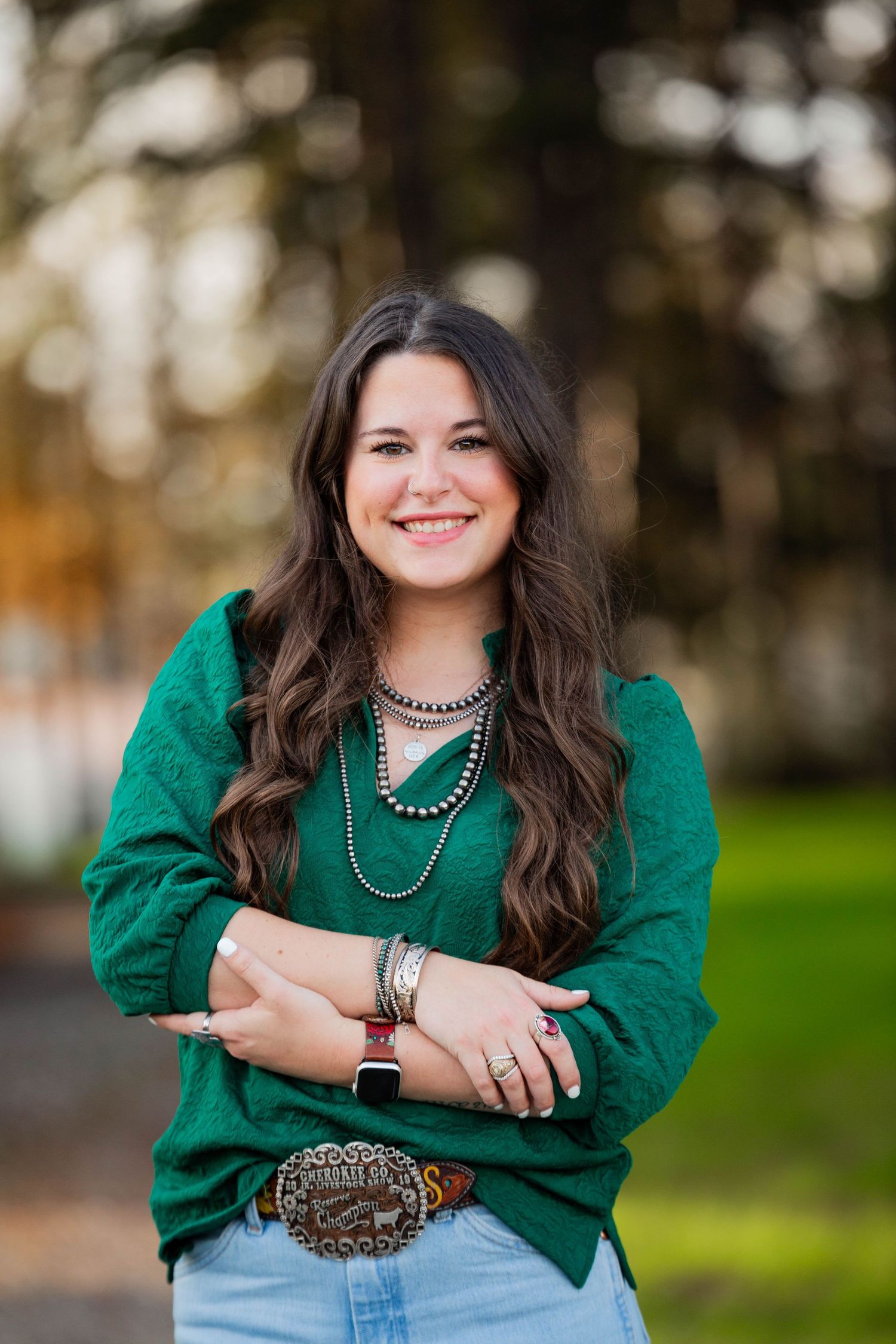 Woman in bright green top and jeans smiles while posing against blurred outdoor background.