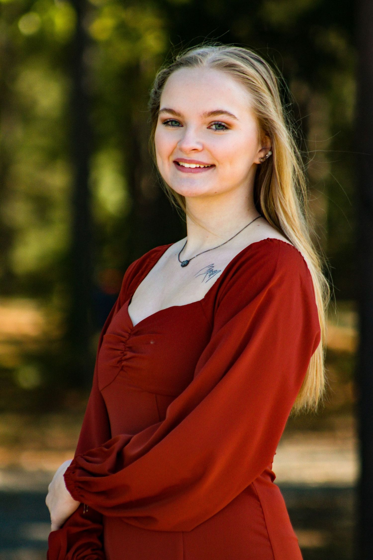 Young blonde woman smiling in a red dress outdoors in a sunlit park setting.