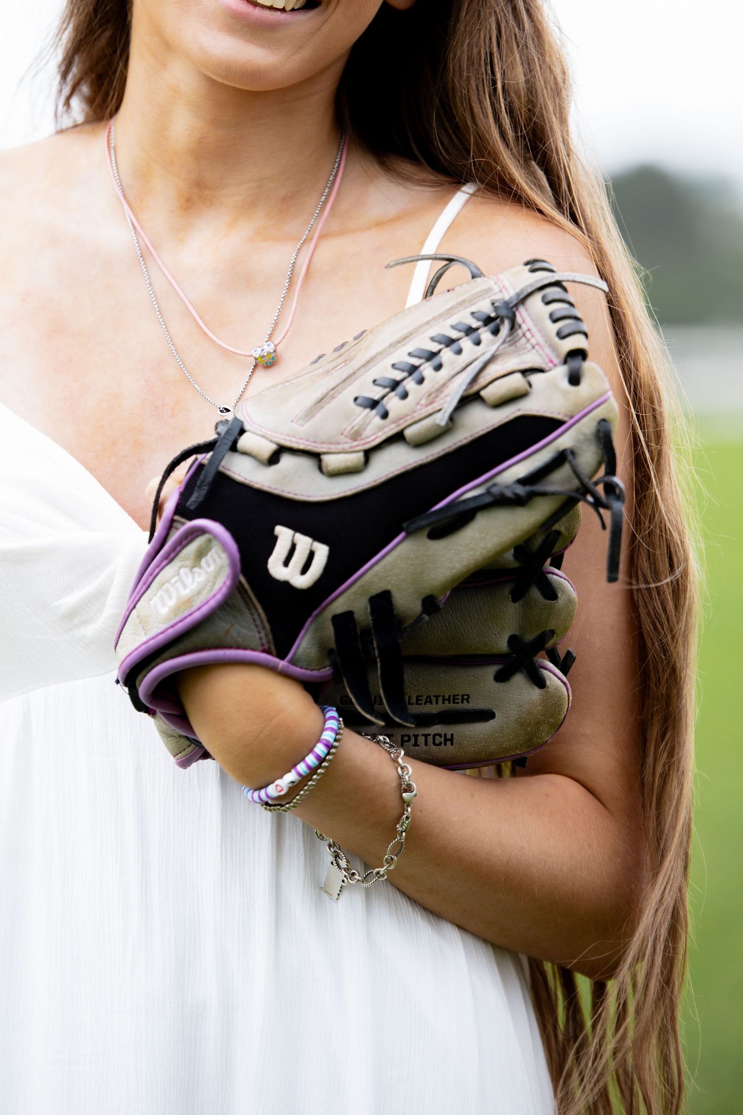 Young woman in white dress holding a Wilson fast pitch softball glove with purple trim outdoors.
