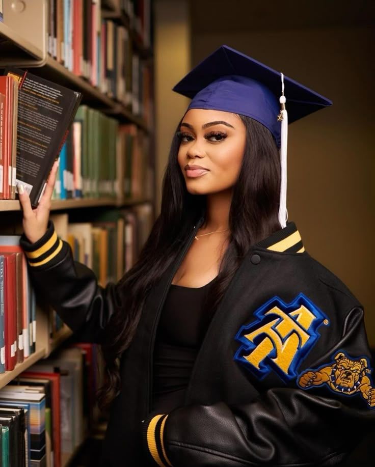 Graduate in cap and gown browses through library books while wearing varsity letterman jacket with blue and gold emblem.